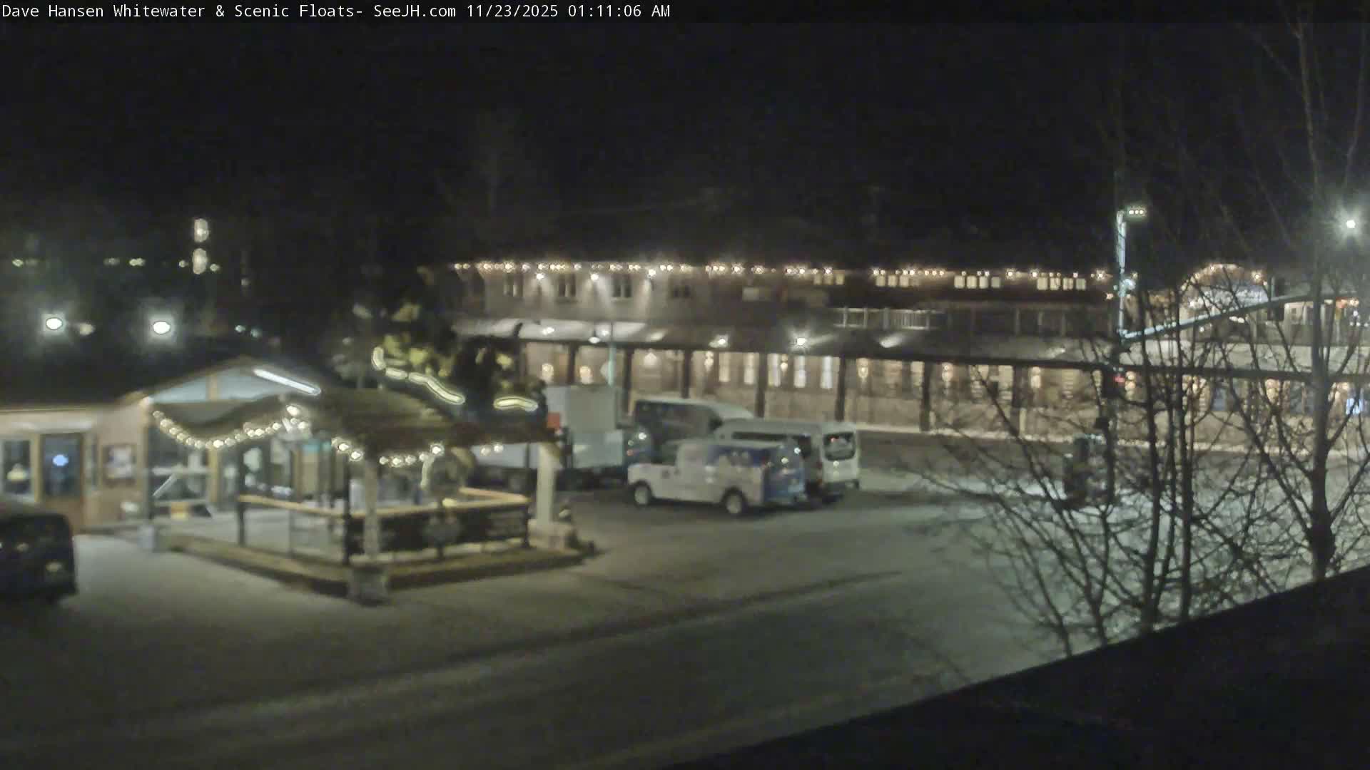 It is a clear night view of an outdoor area featuring several illuminated buildings, one adorned with string lights, and a few parked vehicles on dark ground with bare trees on the right.