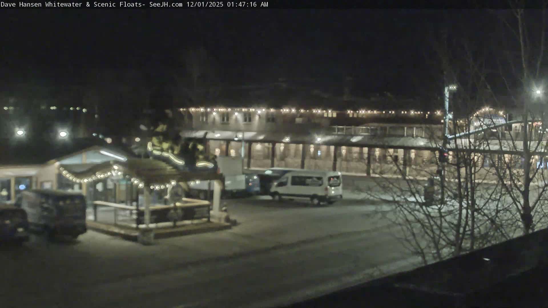 A clear, cold winter night scene depicts illuminated buildings decorated with string lights, a parking lot containing a white van and another vehicle, and bare trees, with patches of snow on the ground.