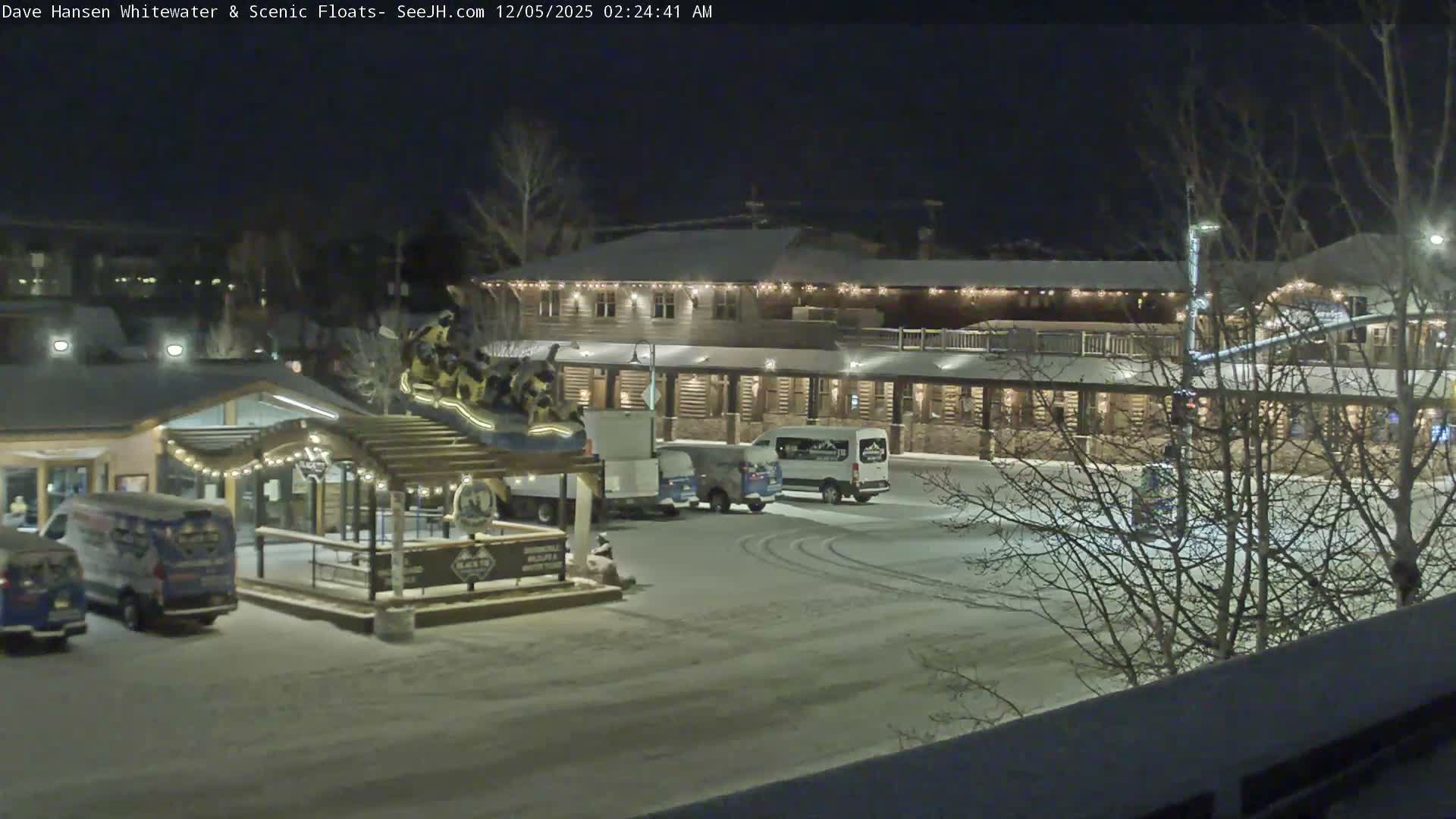 A snow-covered outdoor scene at night features rustic buildings brightly lit with festive string lights, several parked vehicles including vans, bare trees, and a prominent display of yellow rafts, all under clear winter skies.