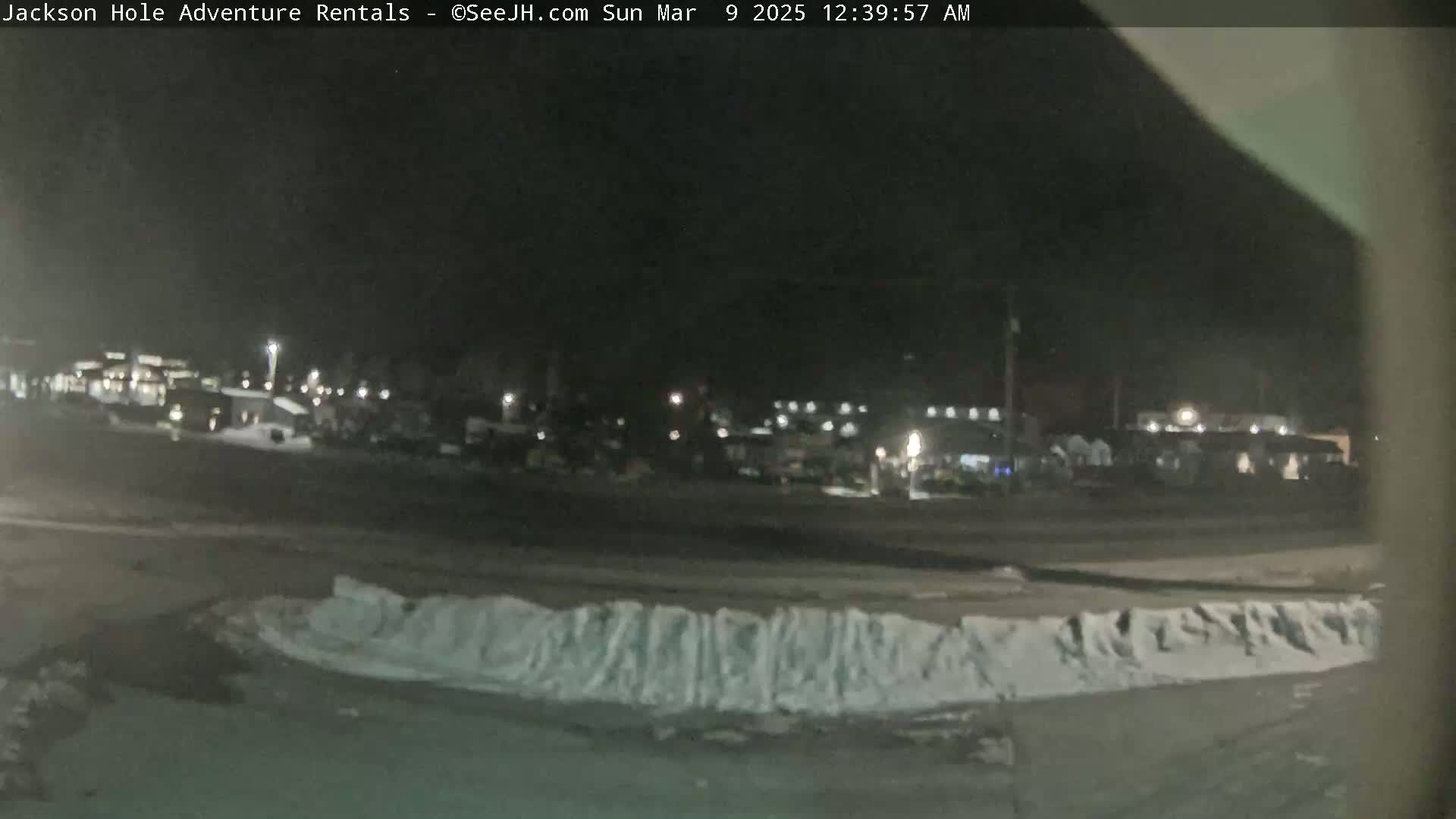 A nighttime, snow-covered parking lot with a row of snow piles in the foreground and dimly lit buildings in the background.