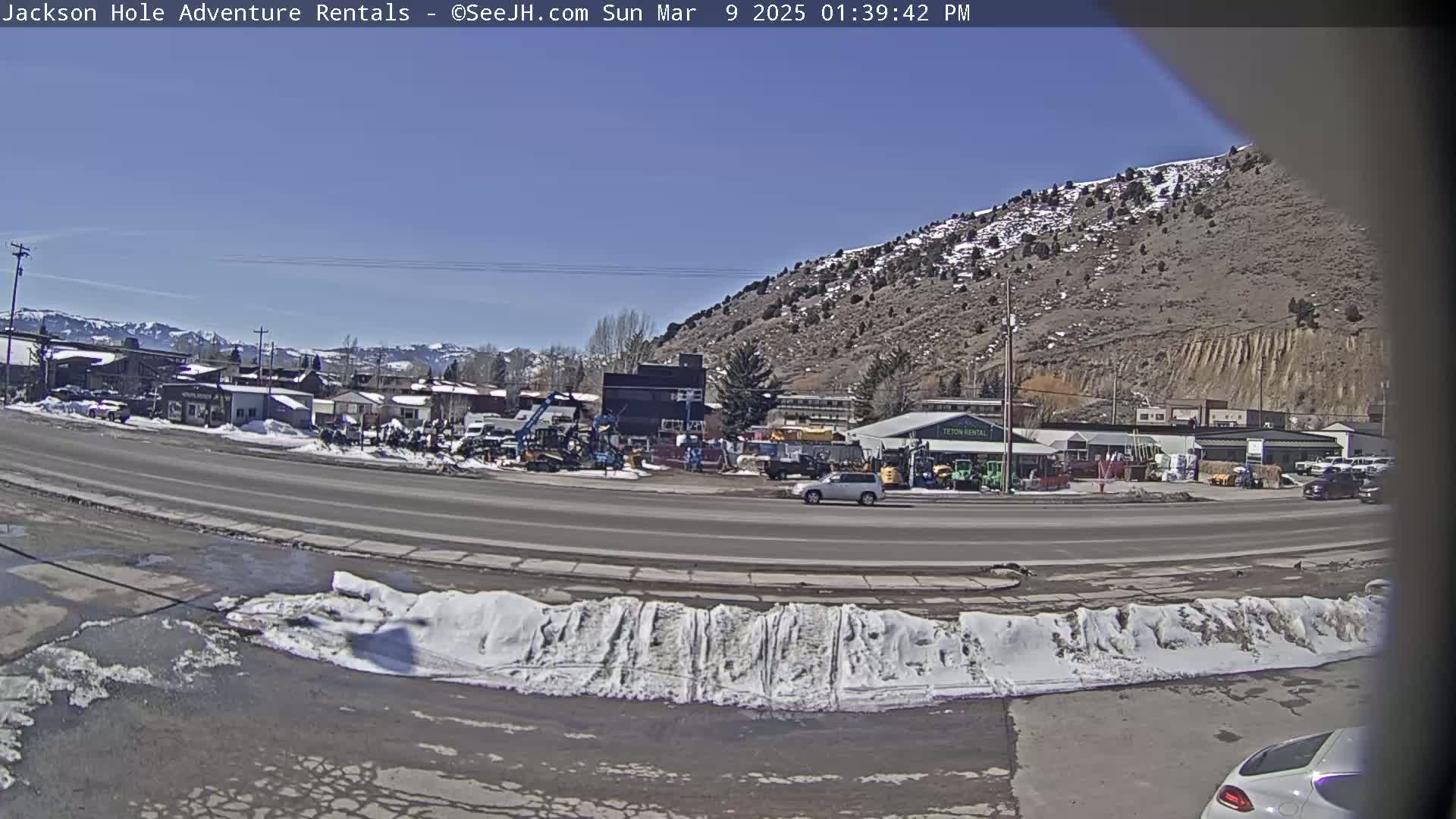 A sunny day reveals a street scene with snow banks along the roadside, featuring a mix of buildings, vehicles, and equipment in front of a mountainous backdrop.