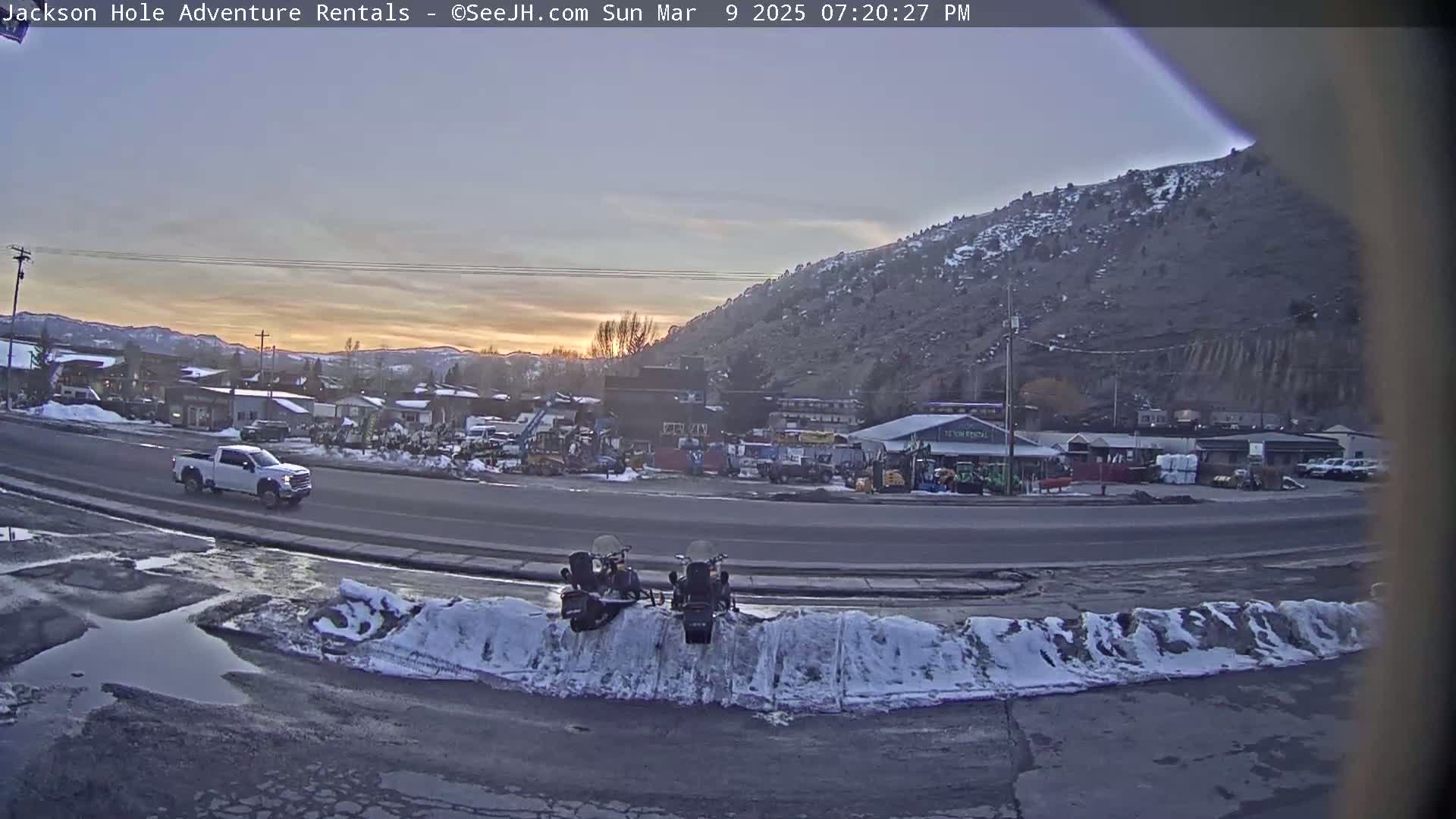 A white pickup truck drives past a snow-covered parking lot with two snowmobiles parked next to a road, overlooking a town at sunset under a mostly clear sky.