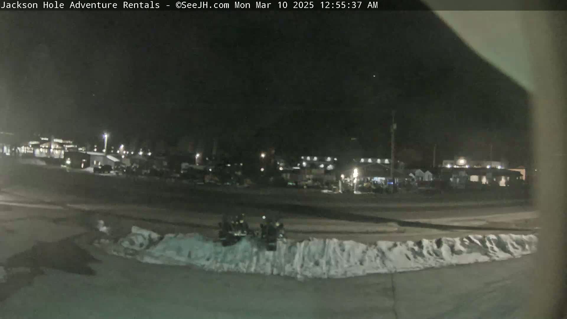 At night, two all-terrain vehicles are parked near a snowdrift in front of a dark, snow-covered parking lot with buildings in the background.