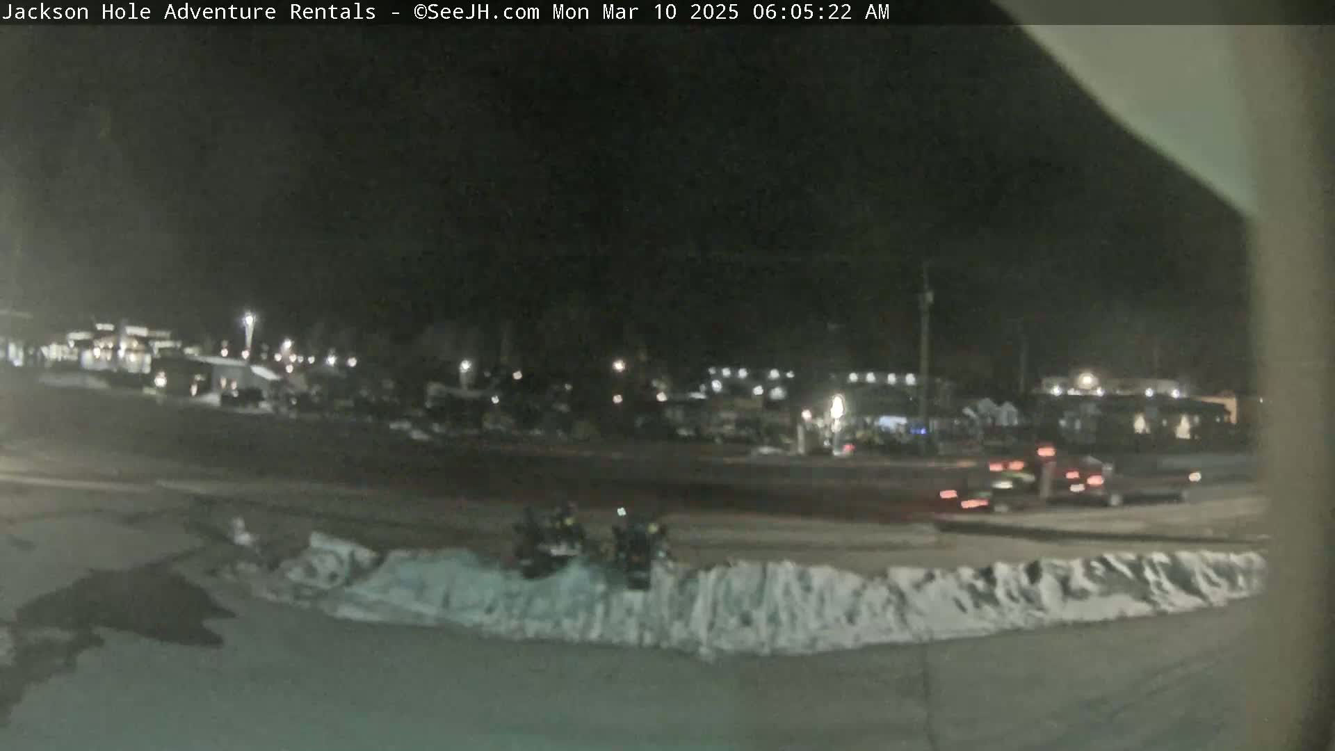 A nighttime, snow-dusted parking lot with several vehicles and two snowmobiles, illuminated by artificial lights.