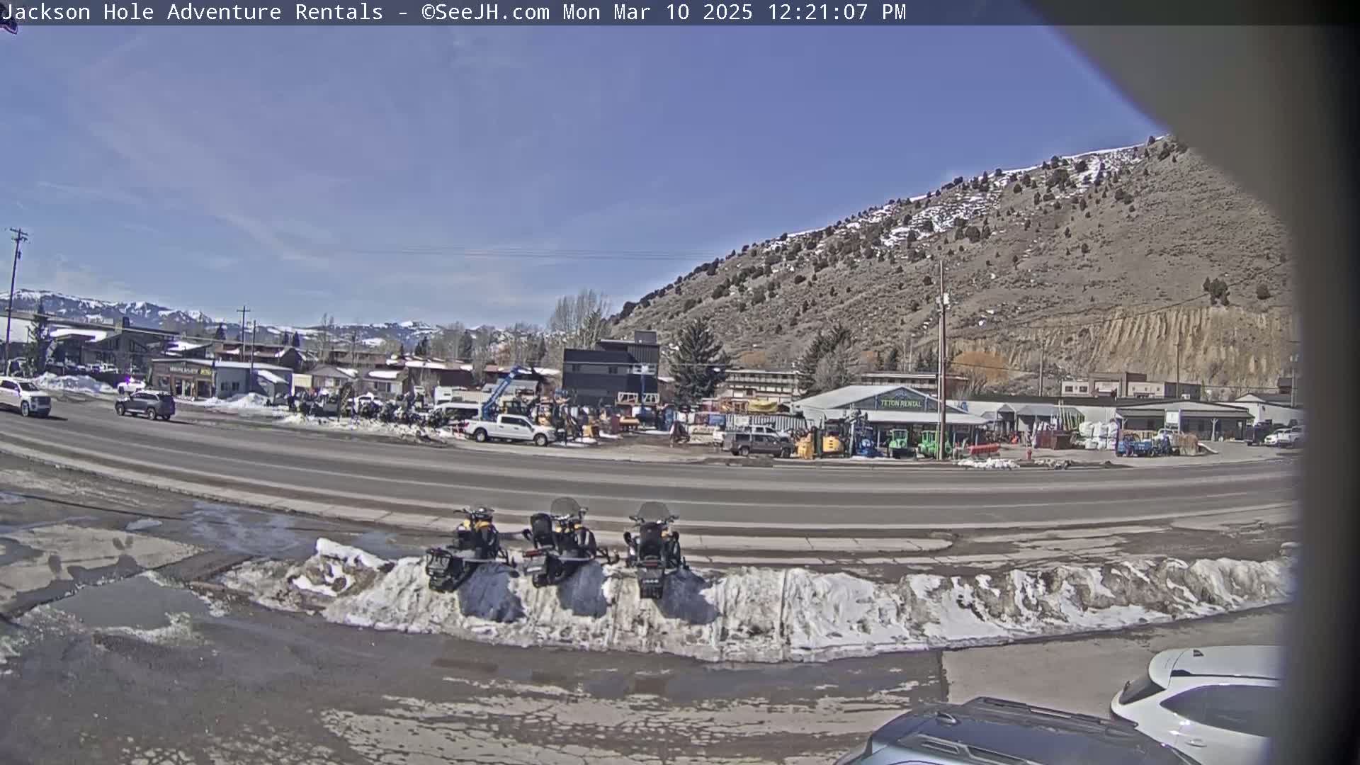 A sunny day reveals a street scene with parked snowmobiles in front of a row of buildings near a mountainside, some of which have patches of snow.