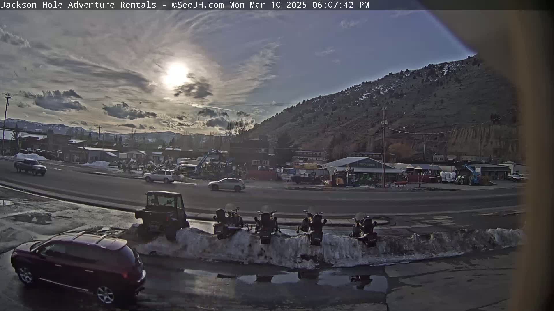 A partly sunny day shows a street scene with several vehicles, including a red SUV, and snow banks lining the road, with a background of buildings and a hill.