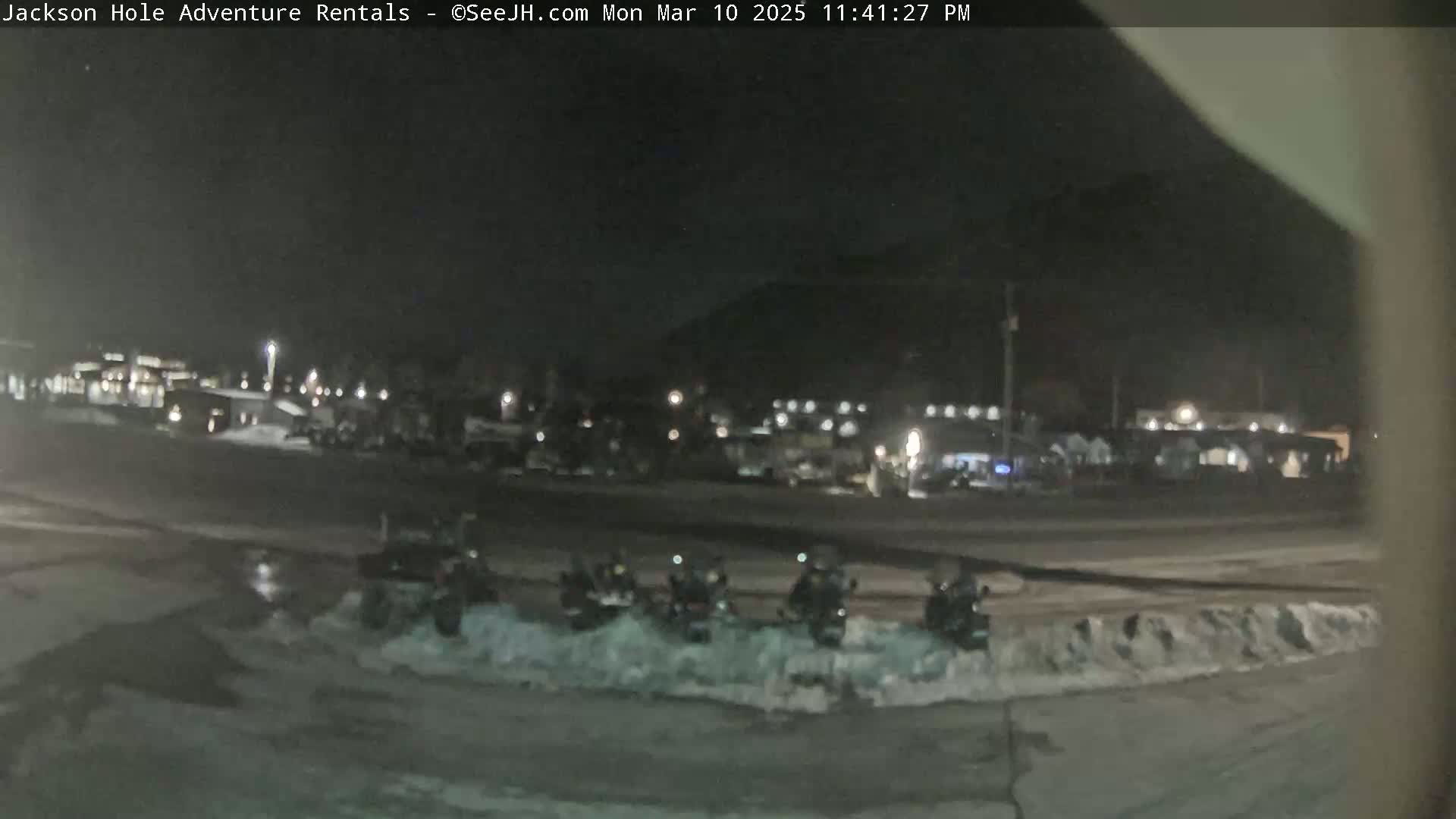 At night, several snowmobiles are parked in a snowy lot in front of a building, under a dark sky.
