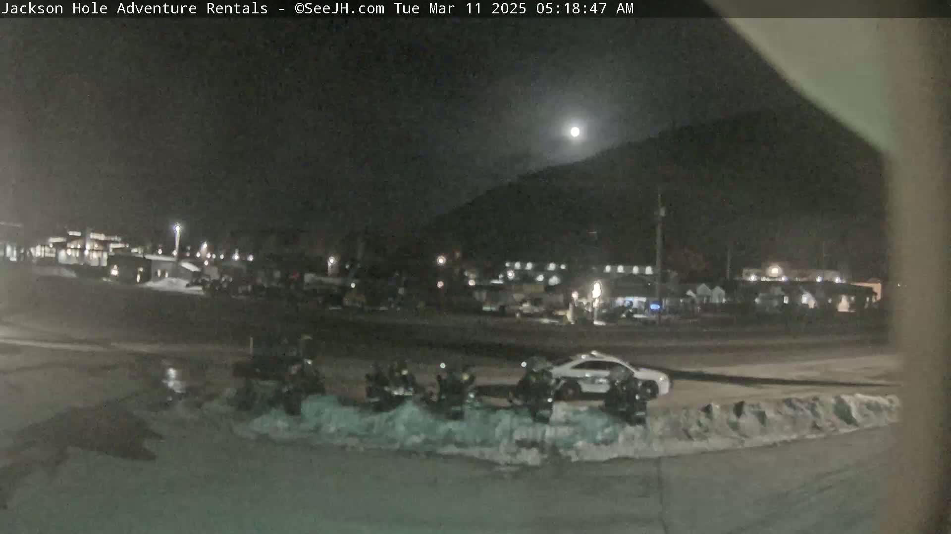 A nighttime scene shows several vehicles parked near snow piles in a parking lot, with a bright moon in the sky and buildings in the background.