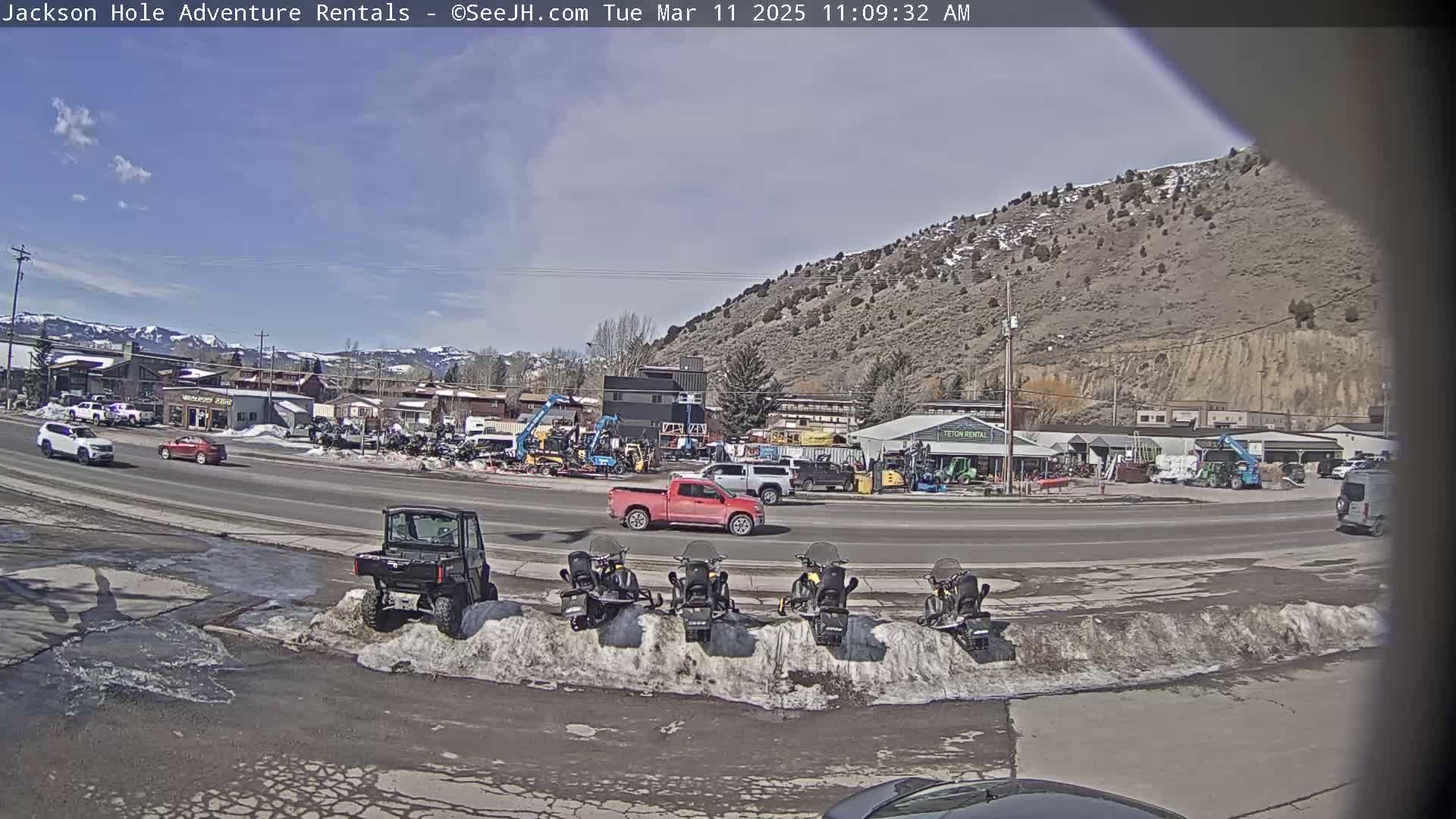 A mostly sunny day reveals a street scene with several vehicles, including a red pickup truck, parked near snow banks and a variety of heavy machinery and snowmobiles at an outdoor rental business, with mountains in the background.