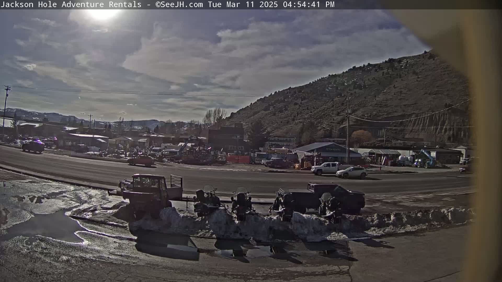 A partly cloudy day shows a road with several vehicles, including snowmobiles parked near a snow bank, in front of a line of buildings nestled at the base of a large, barren hill.