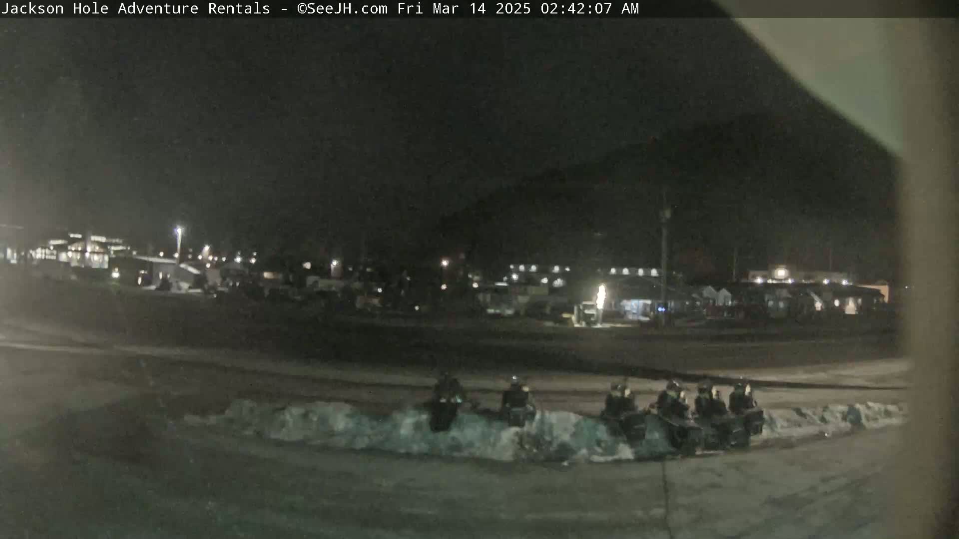 A nighttime scene shows several snowmobiles parked near a snowdrift in front of a building complex under dark, clear skies.