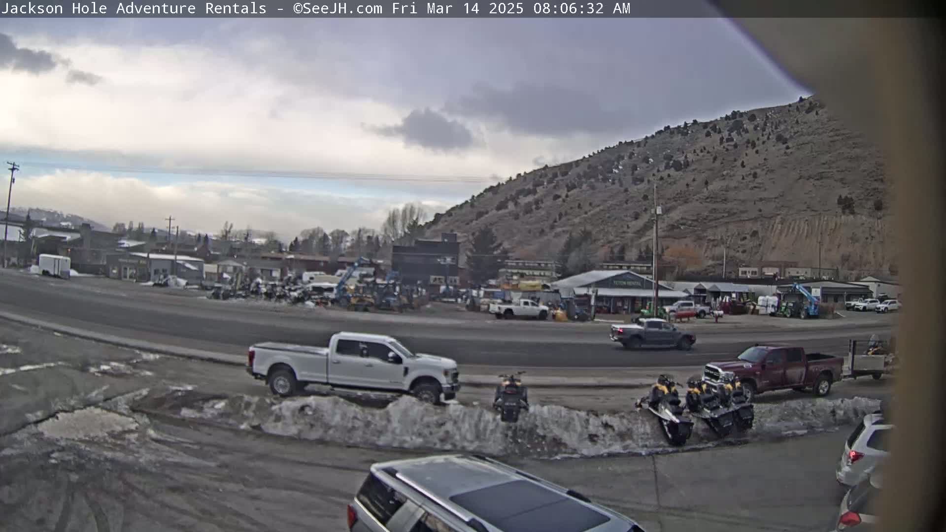 A partly cloudy day reveals a street scene with several vehicles, including trucks and snowmobiles, parked near a row of buildings at the base of a rocky hill.