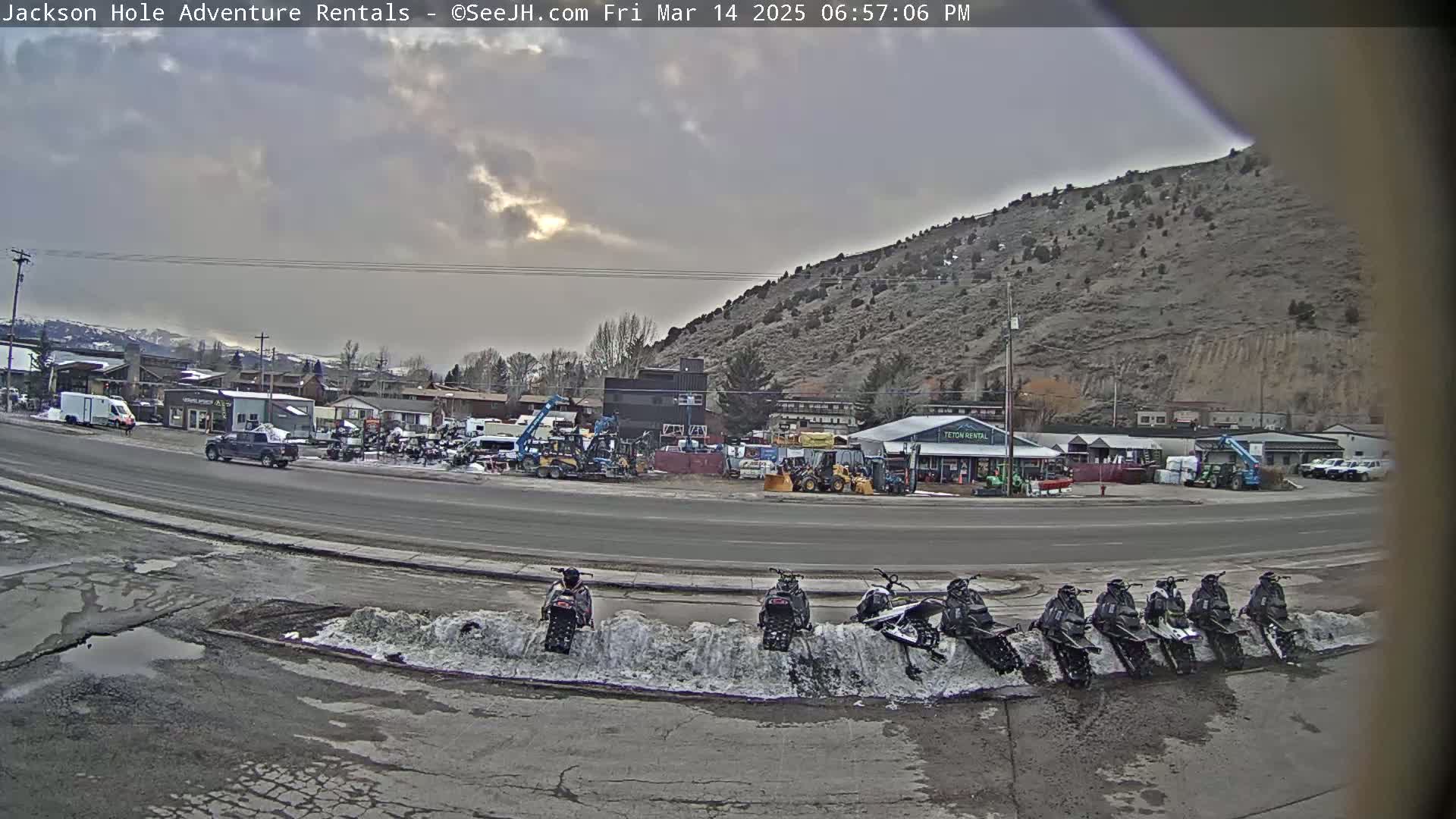 A row of snowmobiles is parked along a curb near a street with several buildings and vehicles under an overcast sky.