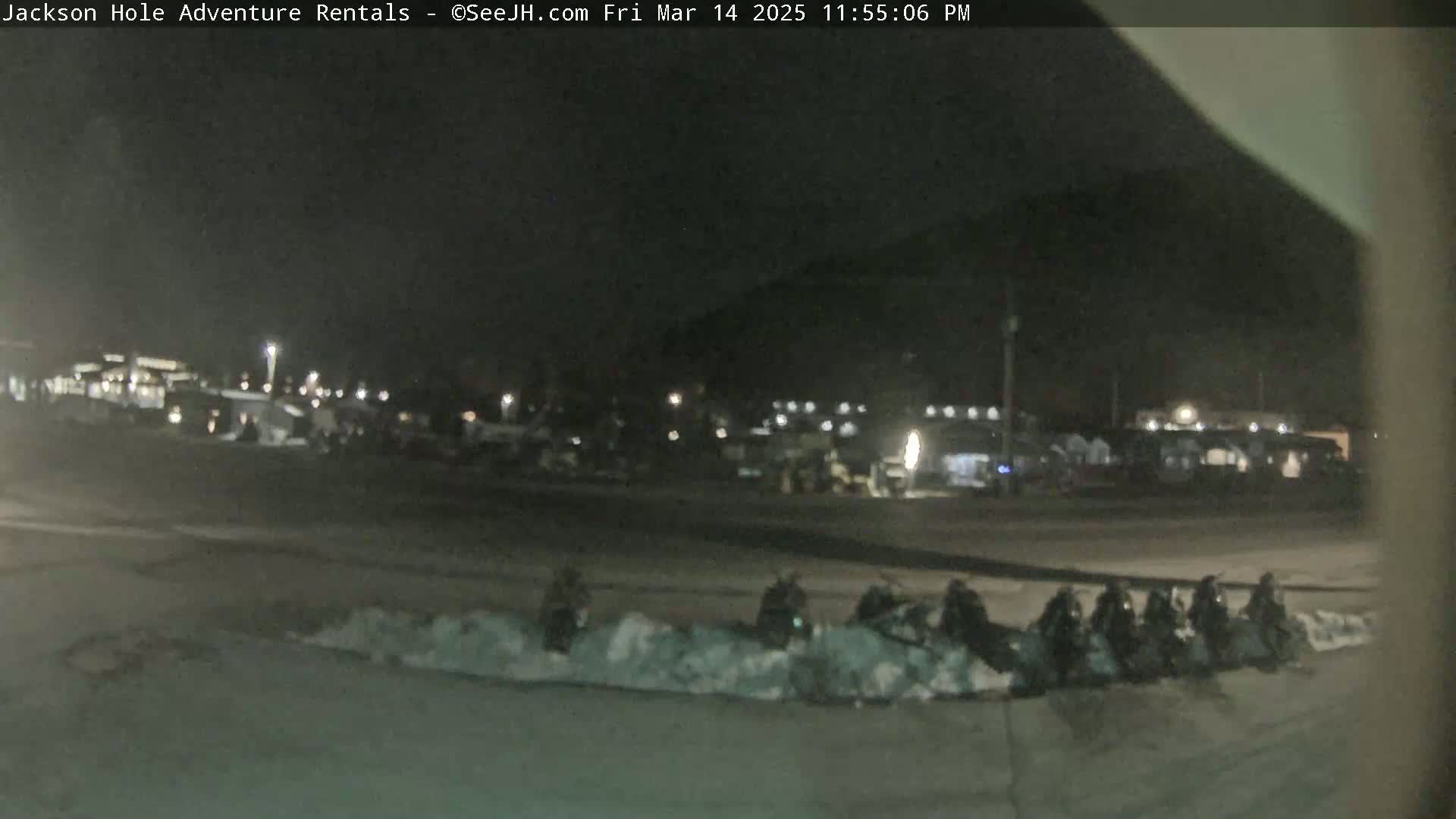 At night, several snowmobiles are parked in a snowy area near buildings under dim lighting.