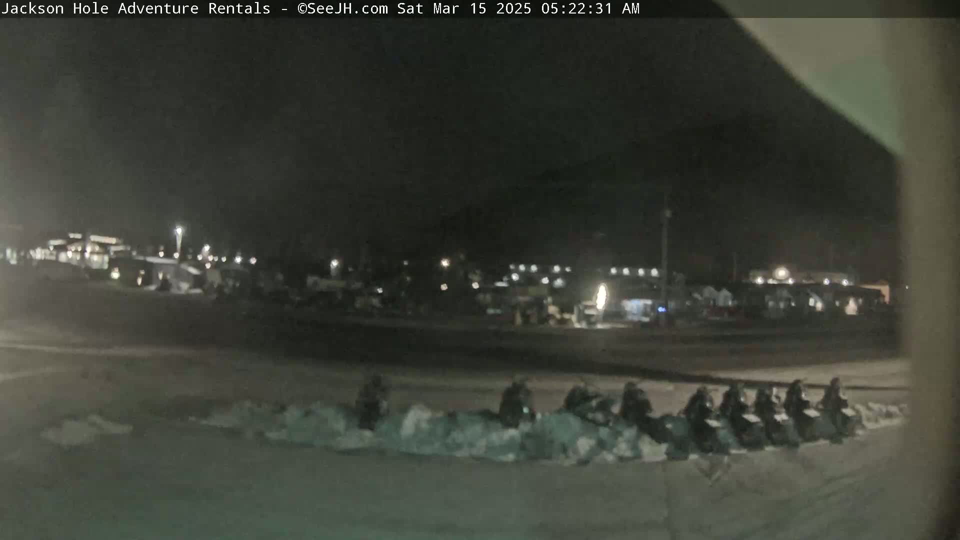 A nighttime, snowy scene shows a line of snowmobiles parked in a snow-covered area in front of a dimly lit building.
