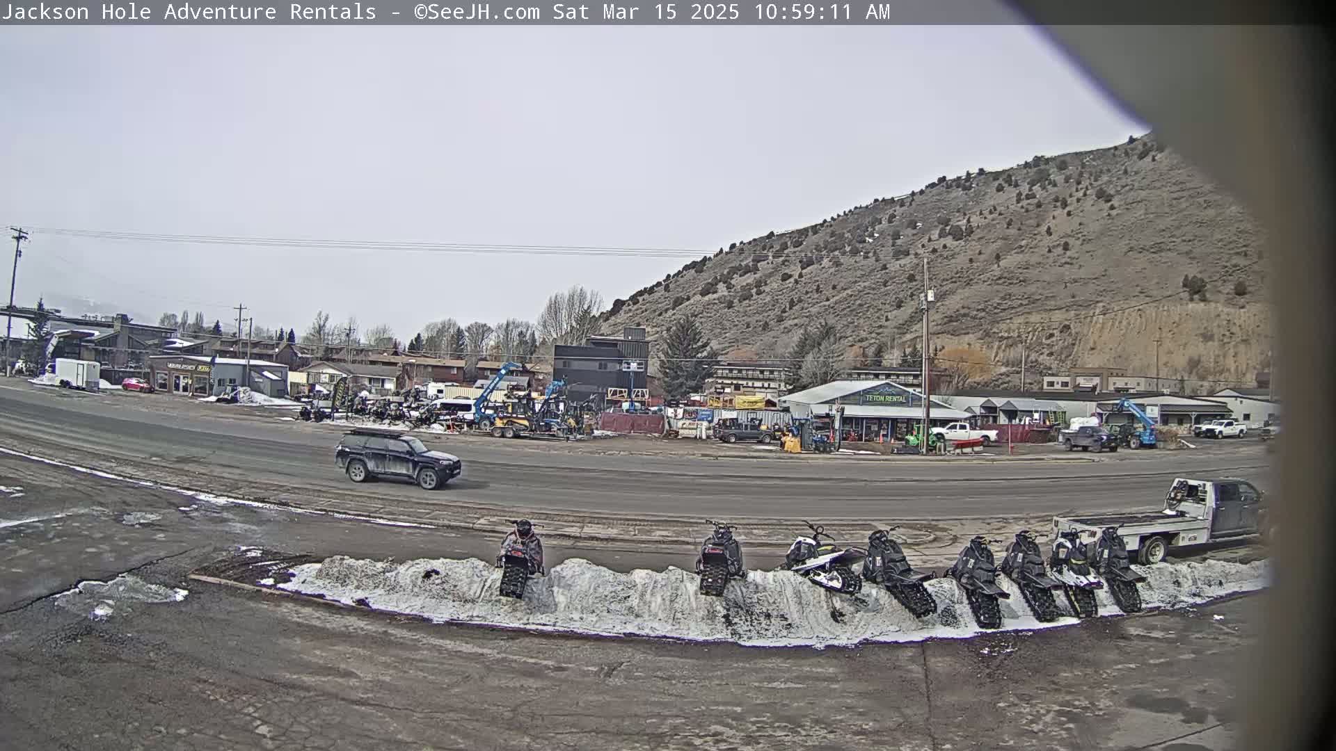 A street scene on an overcast day shows several snowmobiles parked in a row against a snowbank, a dark-colored SUV driving on a road in front of various buildings and equipment, and a flatbed truck further down the road.