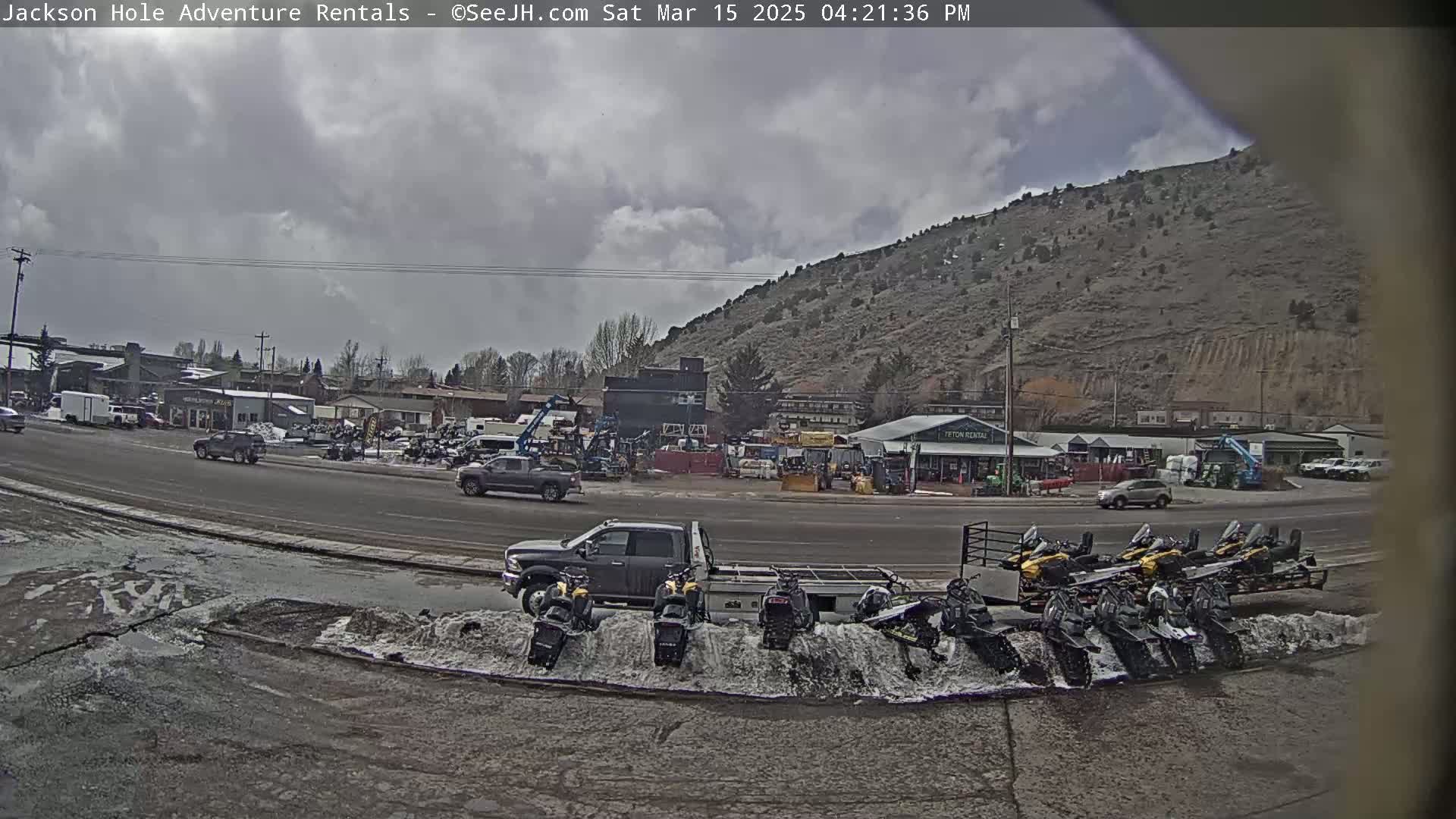 A cloudy day reveals a parking lot full of snowmobiles and other vehicles near a hill, with several buildings and some cars in the background.