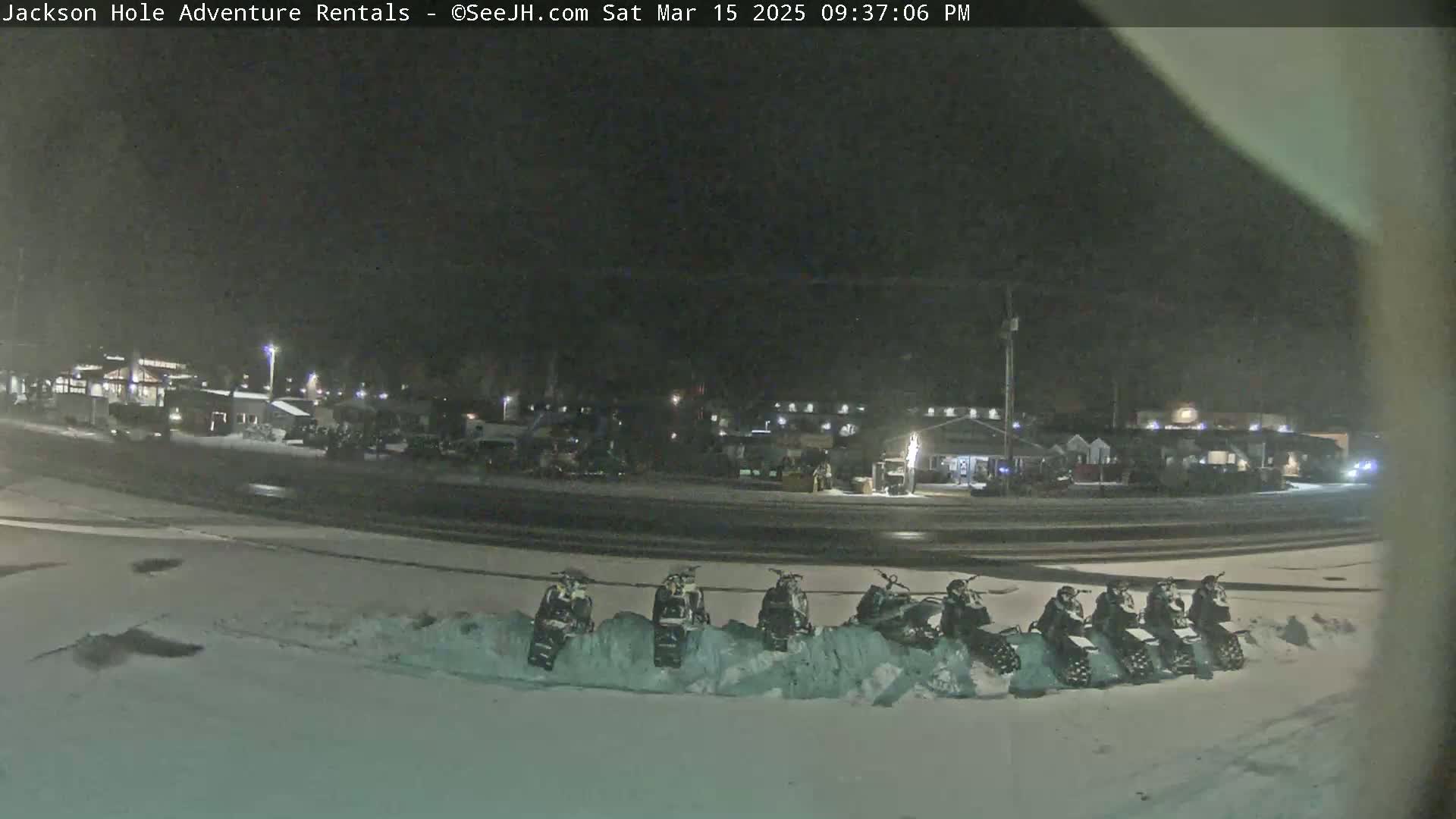 A nighttime, snow-covered parking lot contains several snowmobiles parked in a row near a road and some buildings.