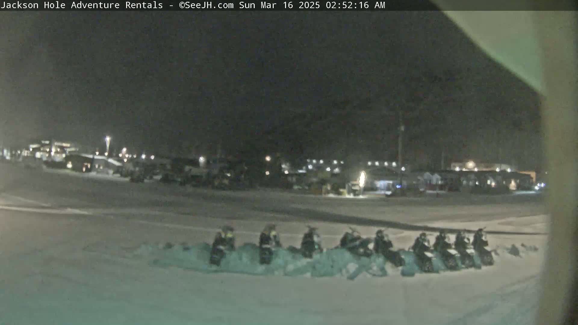 A nighttime, snowy scene shows a row of snowmobiles parked in a snowdrift in front of a building complex.