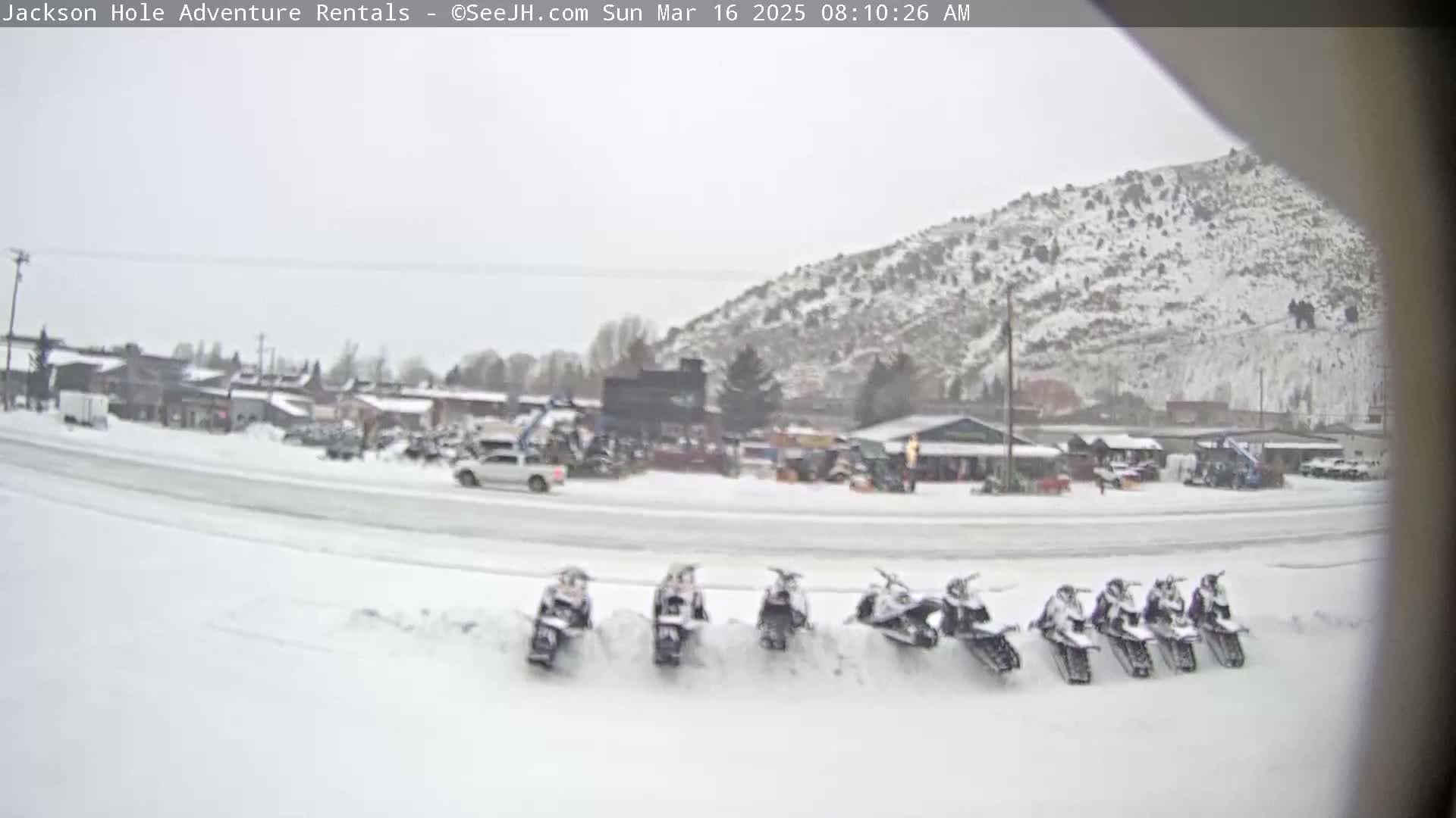 A snow-covered town with a row of snowmobiles parked in front of some buildings, with a snow-covered mountain in the background under overcast skies.