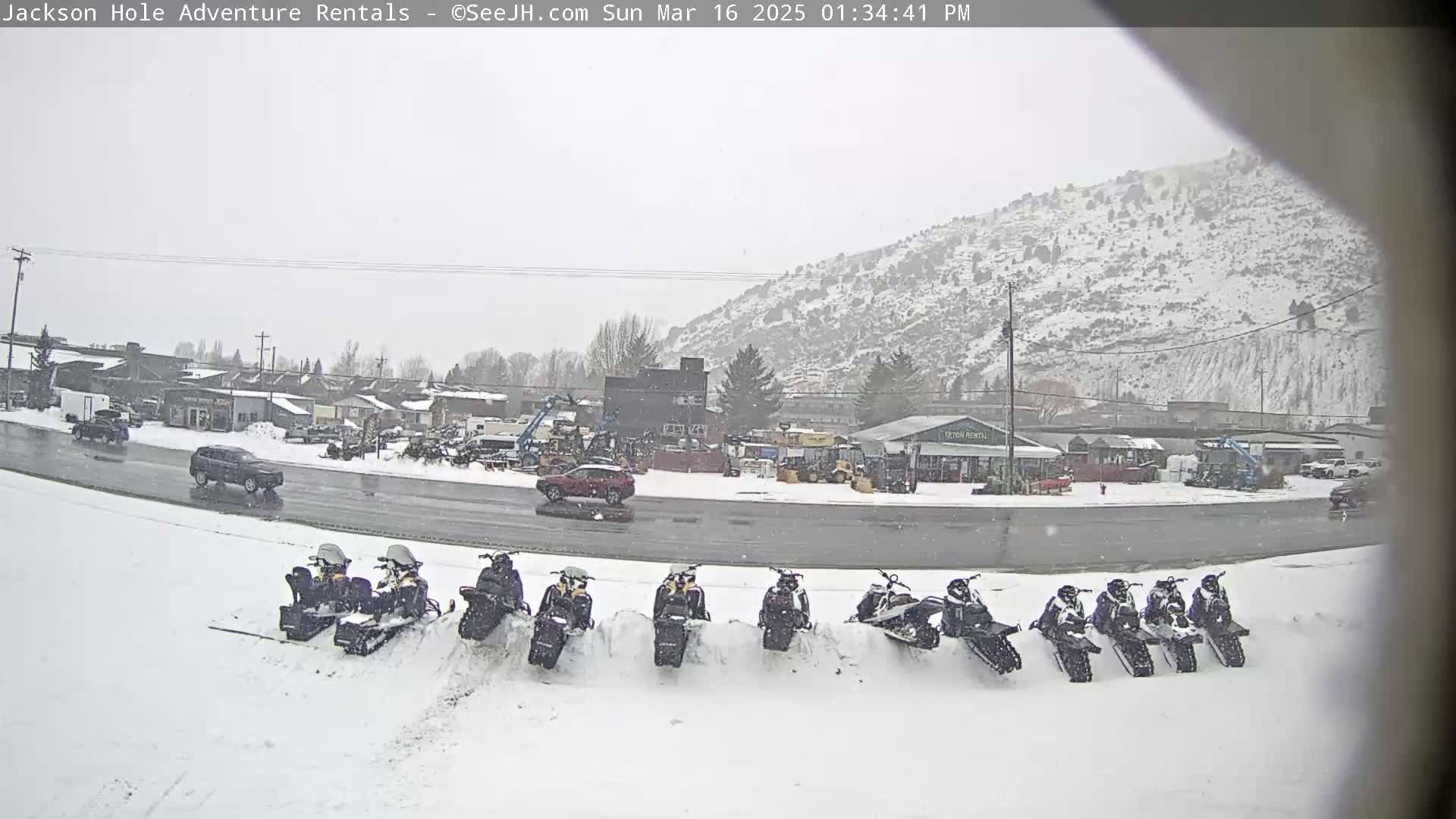 A row of snowmobiles sits in the snow in front of a light-snow-covered road and buildings, with a snow-covered mountain in the background.