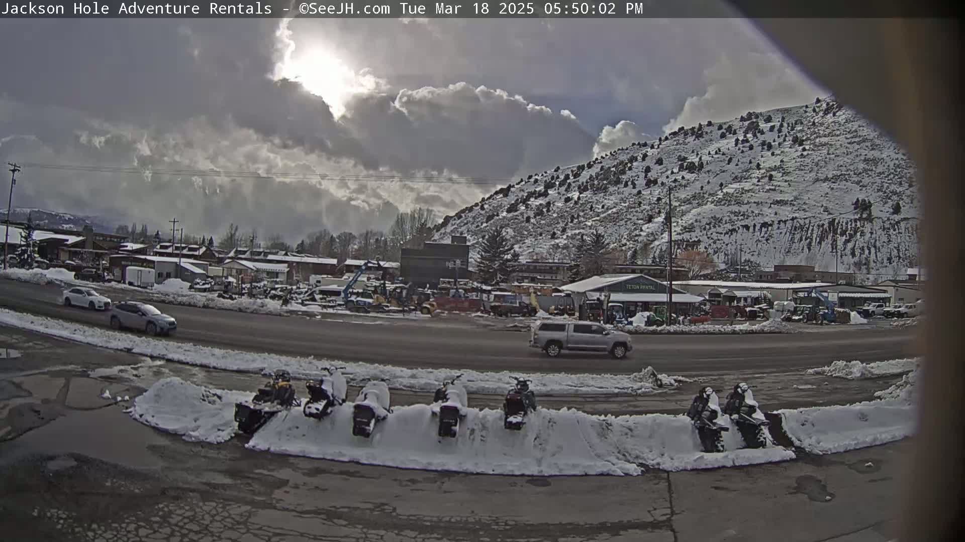 A snow-covered town sits beneath a partly cloudy sky, with several snowmobiles parked in a row along a snow bank next to a road.
