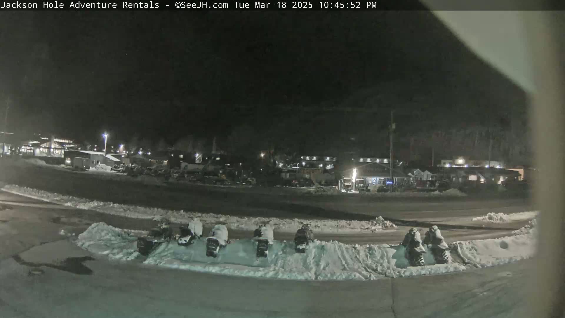 A nighttime view of a snow-covered parking lot with several snowmobiles parked, in front of a cluster of buildings under a dark sky.