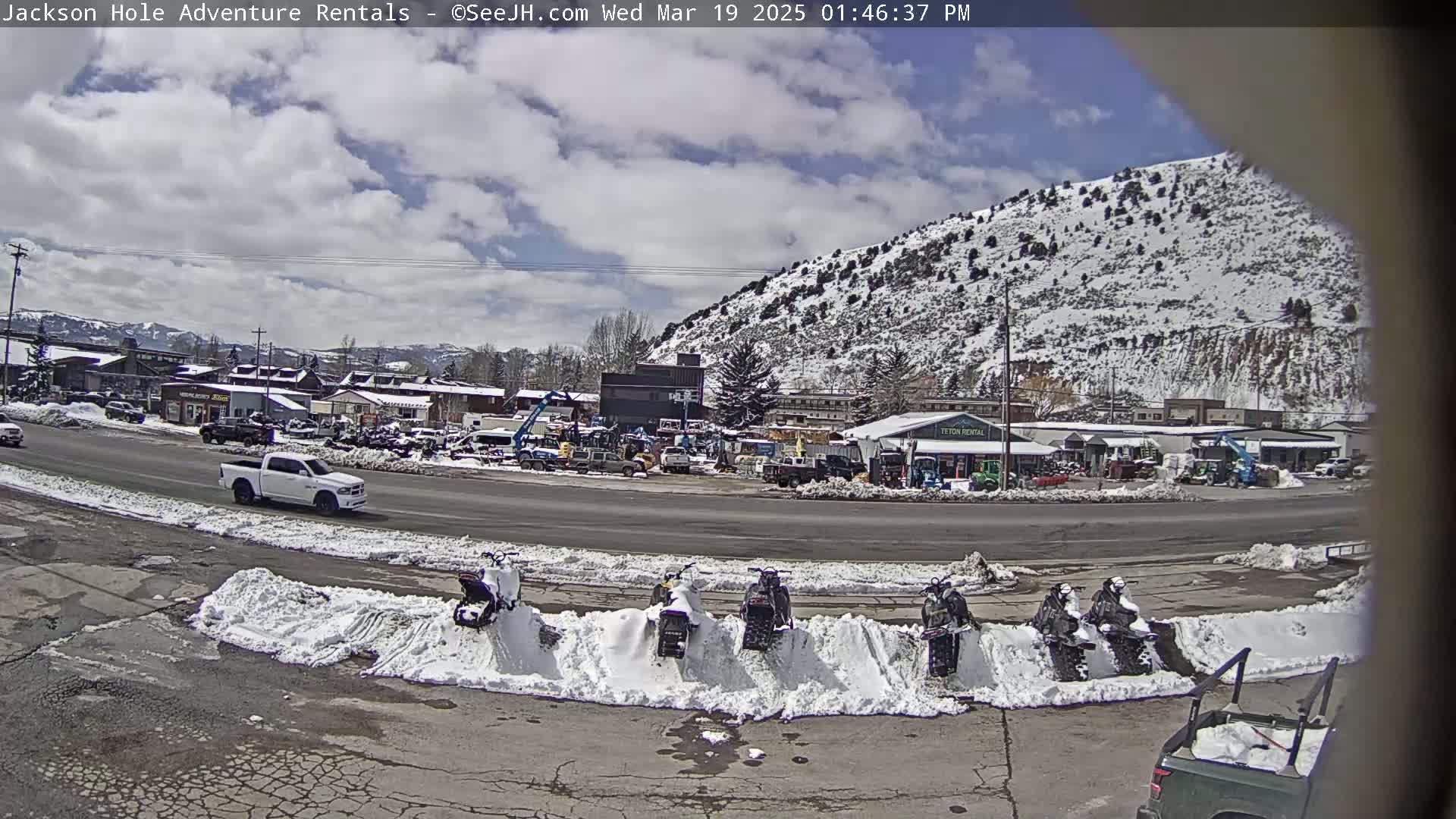 A partly cloudy day shows a snow-covered mountain behind a street scene with a white pickup truck, several snowmobiles parked in the snow, and various vehicles and equipment at a rental business.