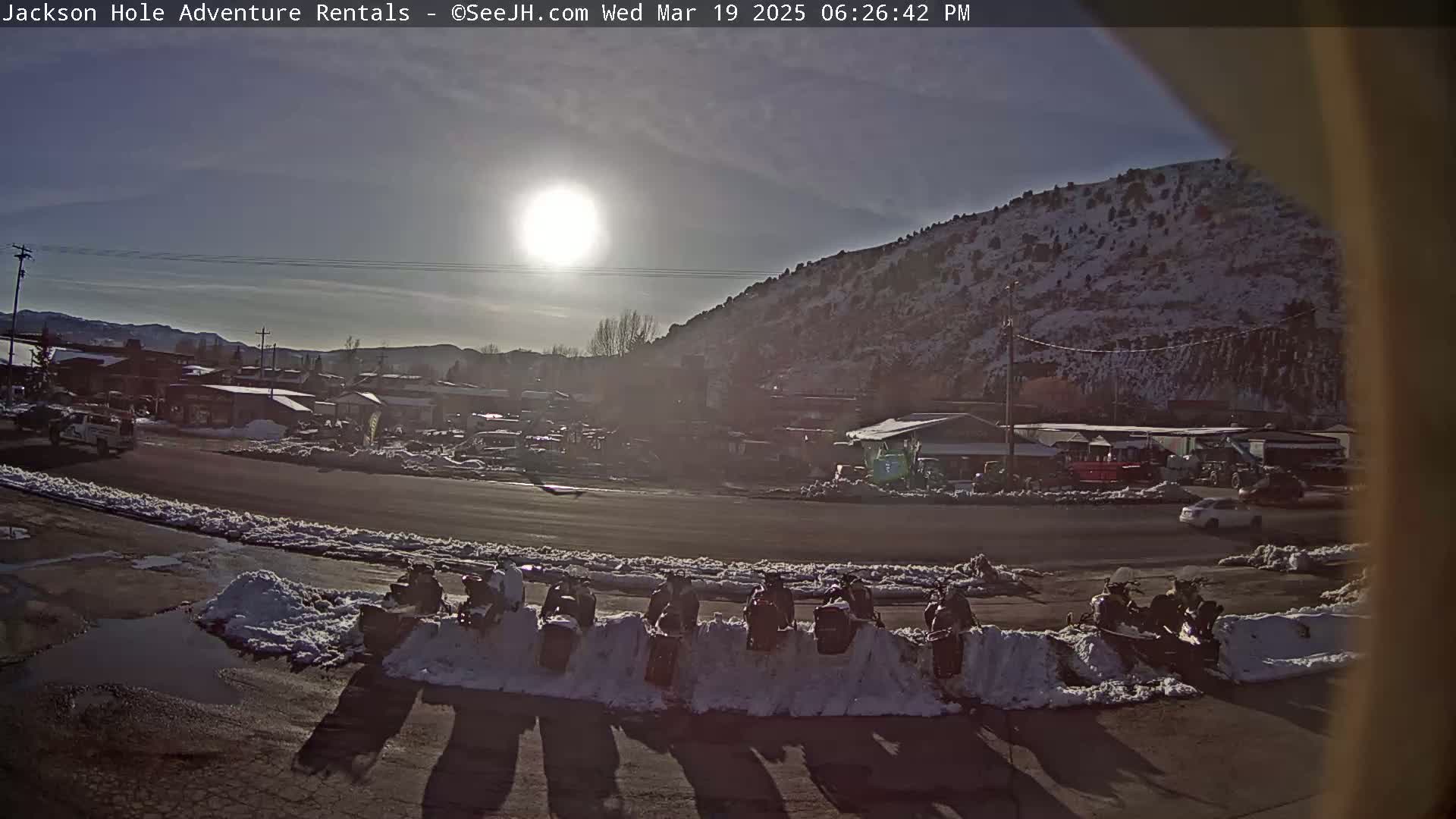 A sunny day reveals a snow-covered parking lot with several snowmobiles lined up, near a town nestled at the base of a snow-dusted mountain.