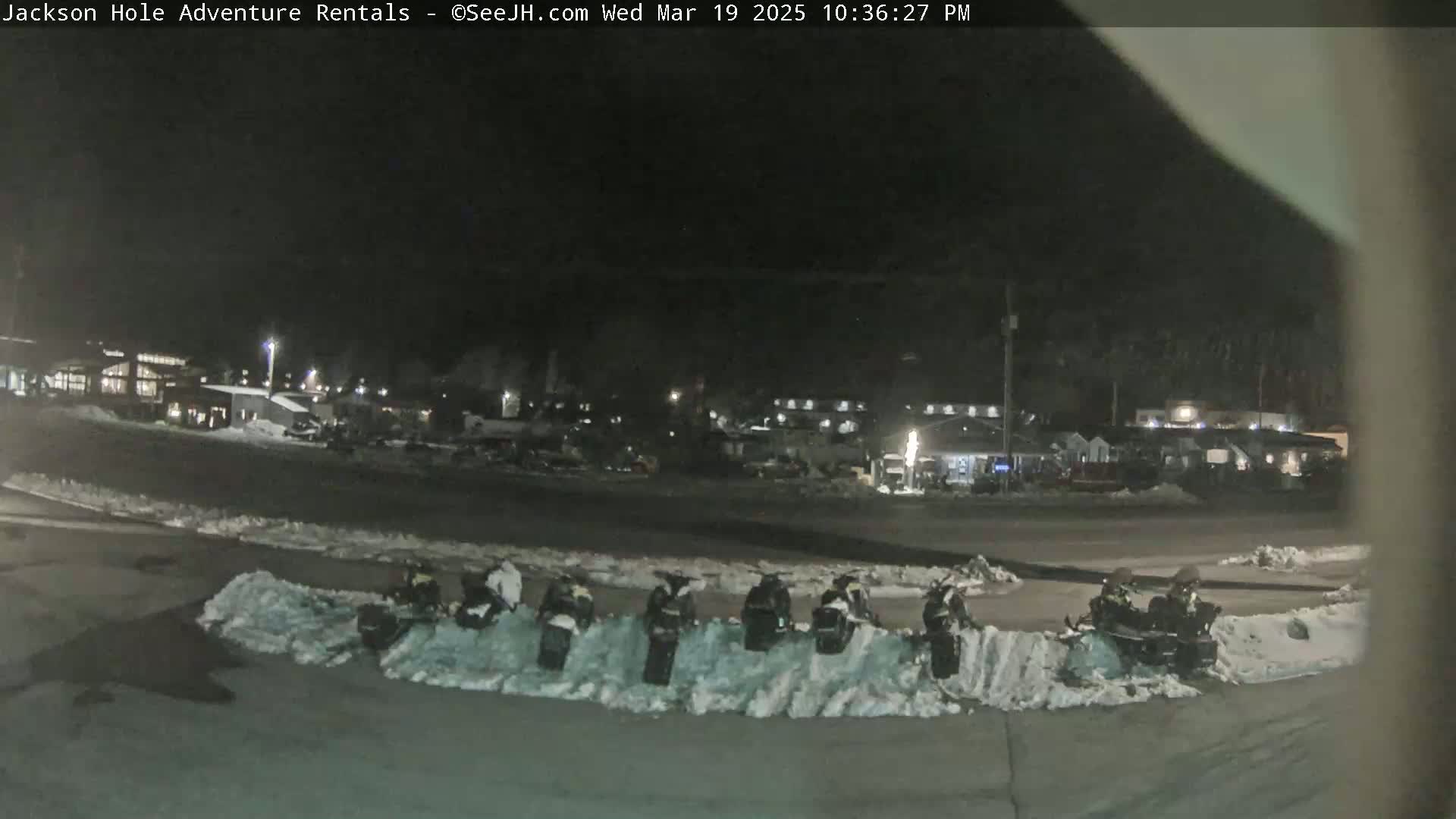 At night, several snowmobiles are parked in a snow-covered lot in front of a building complex.
