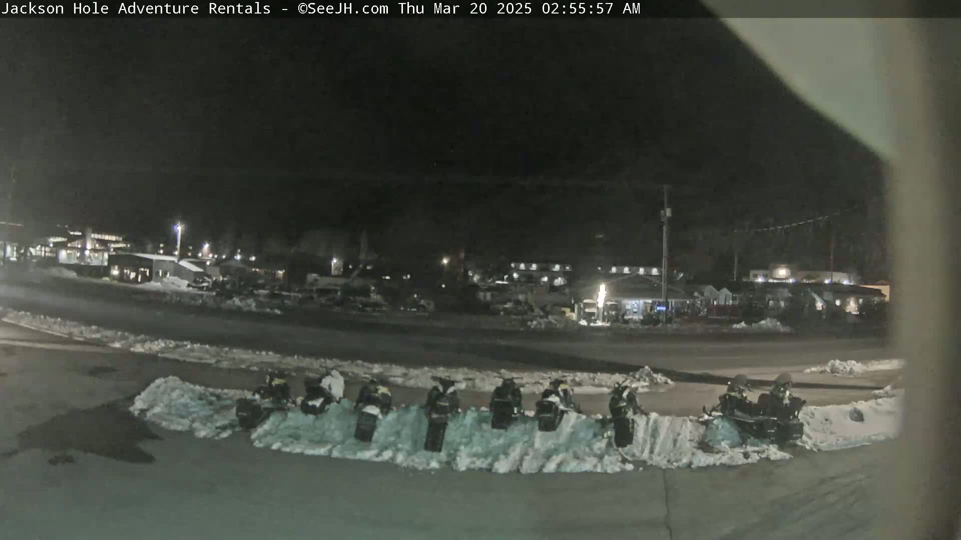Several snowmobiles are parked in a snow-covered lot at night, with a background of buildings and a dark sky.