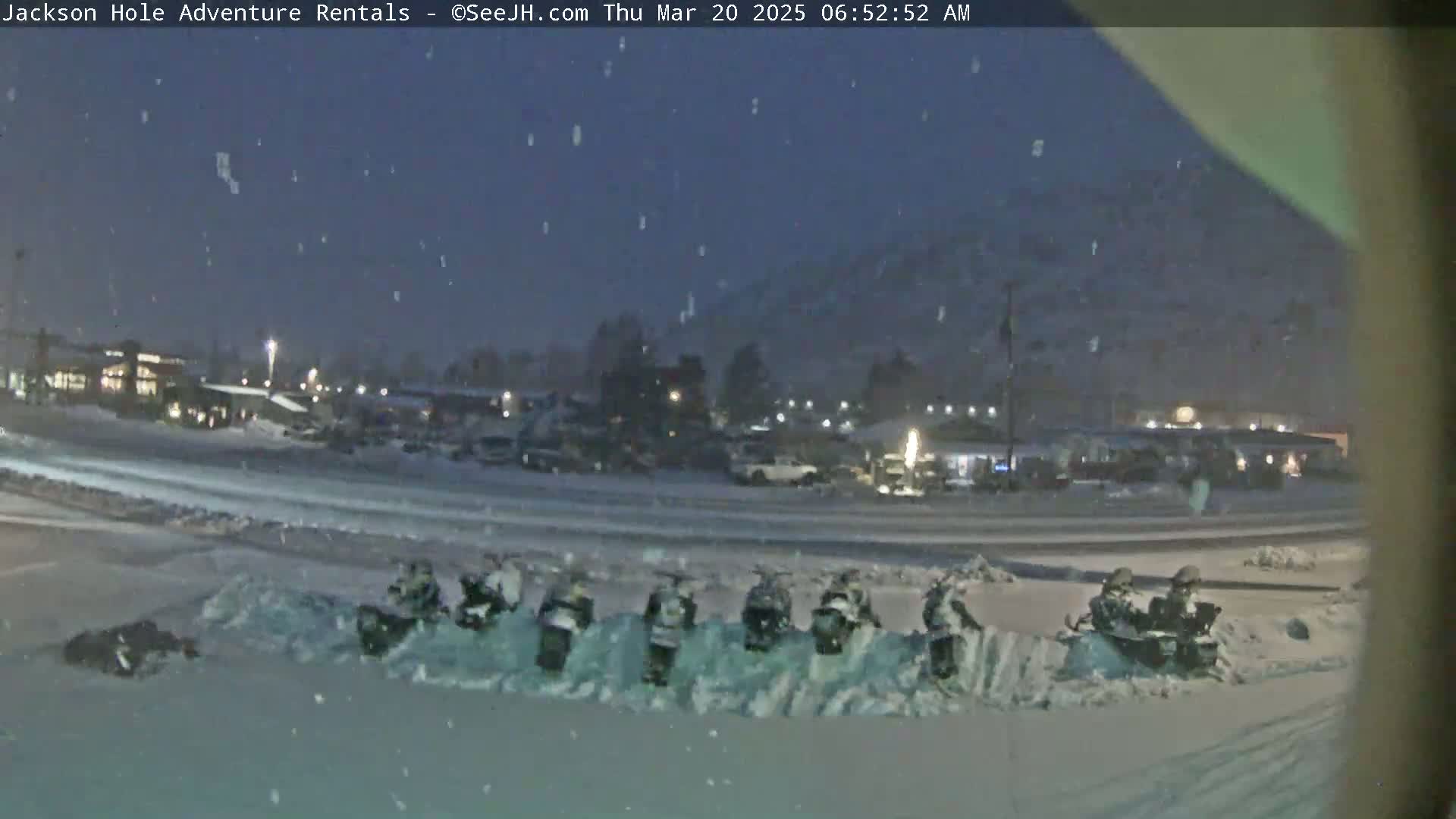A snow-covered parking lot at night, with several snowmobiles parked in a row, under light snowfall.