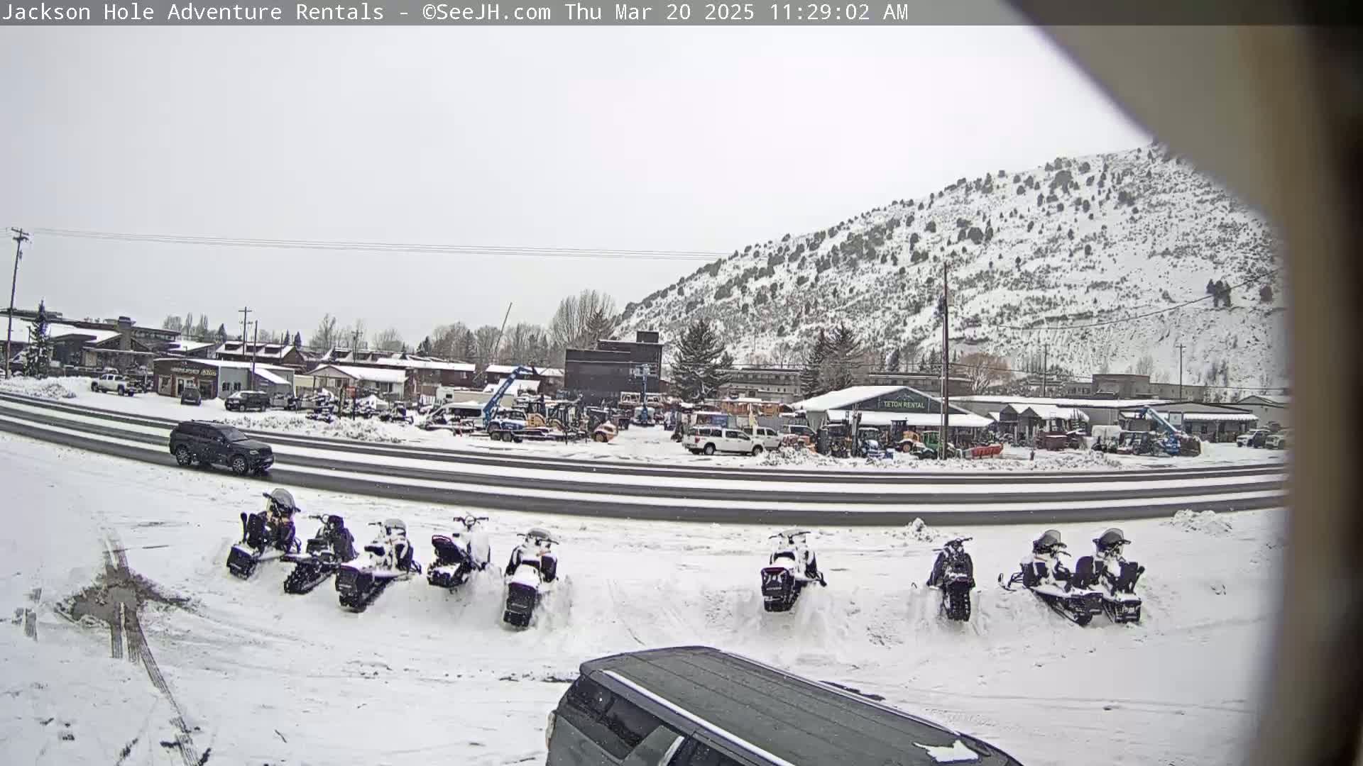 A snow-covered parking lot with several snowmobiles parked in the snow, in front of a road and buildings with a snow-covered hillside in the background under an overcast sky.