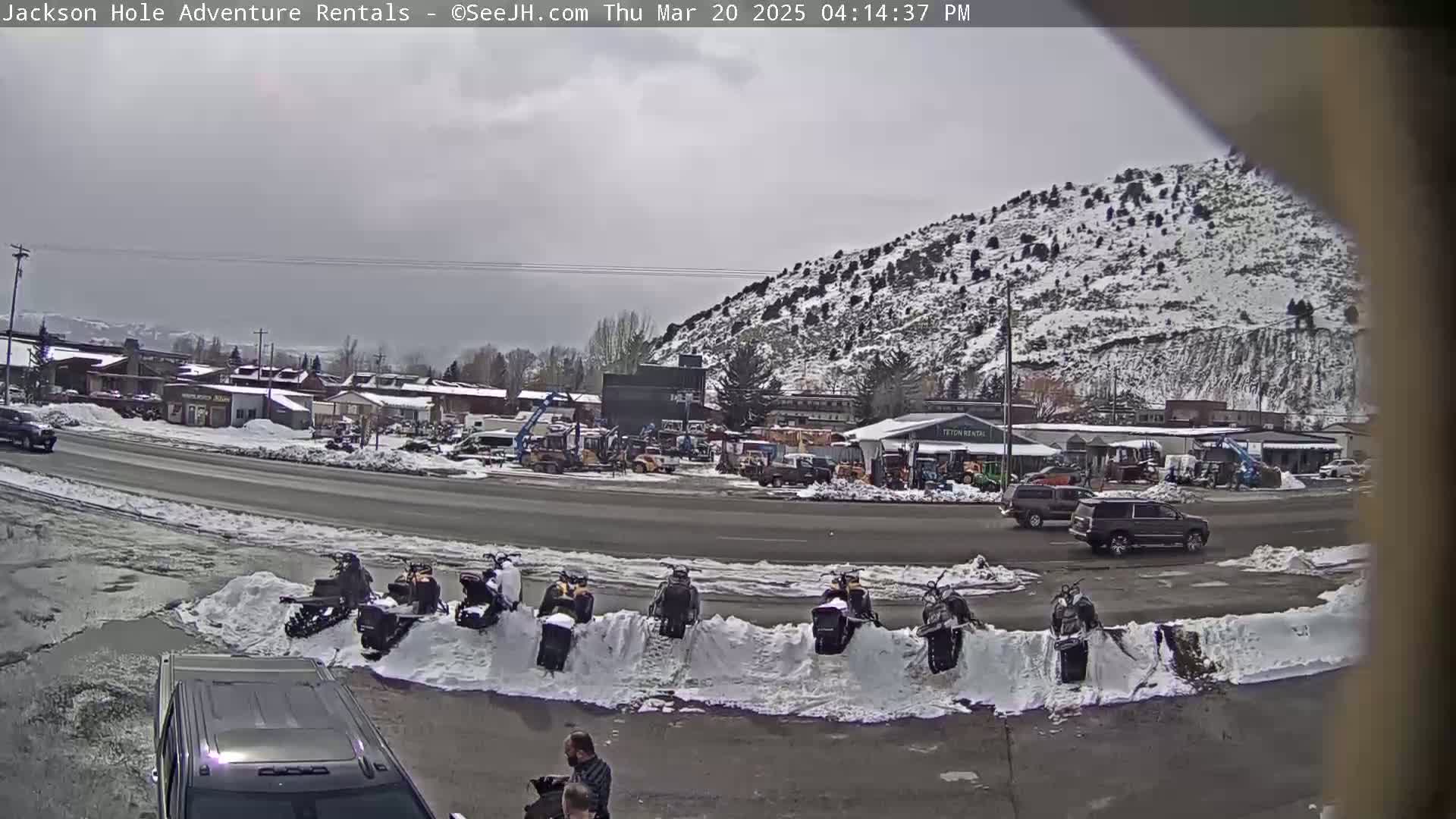 A snow-covered hill overlooks a street scene with several snowmobiles parked in the snow, some cars, and buildings under an overcast sky.