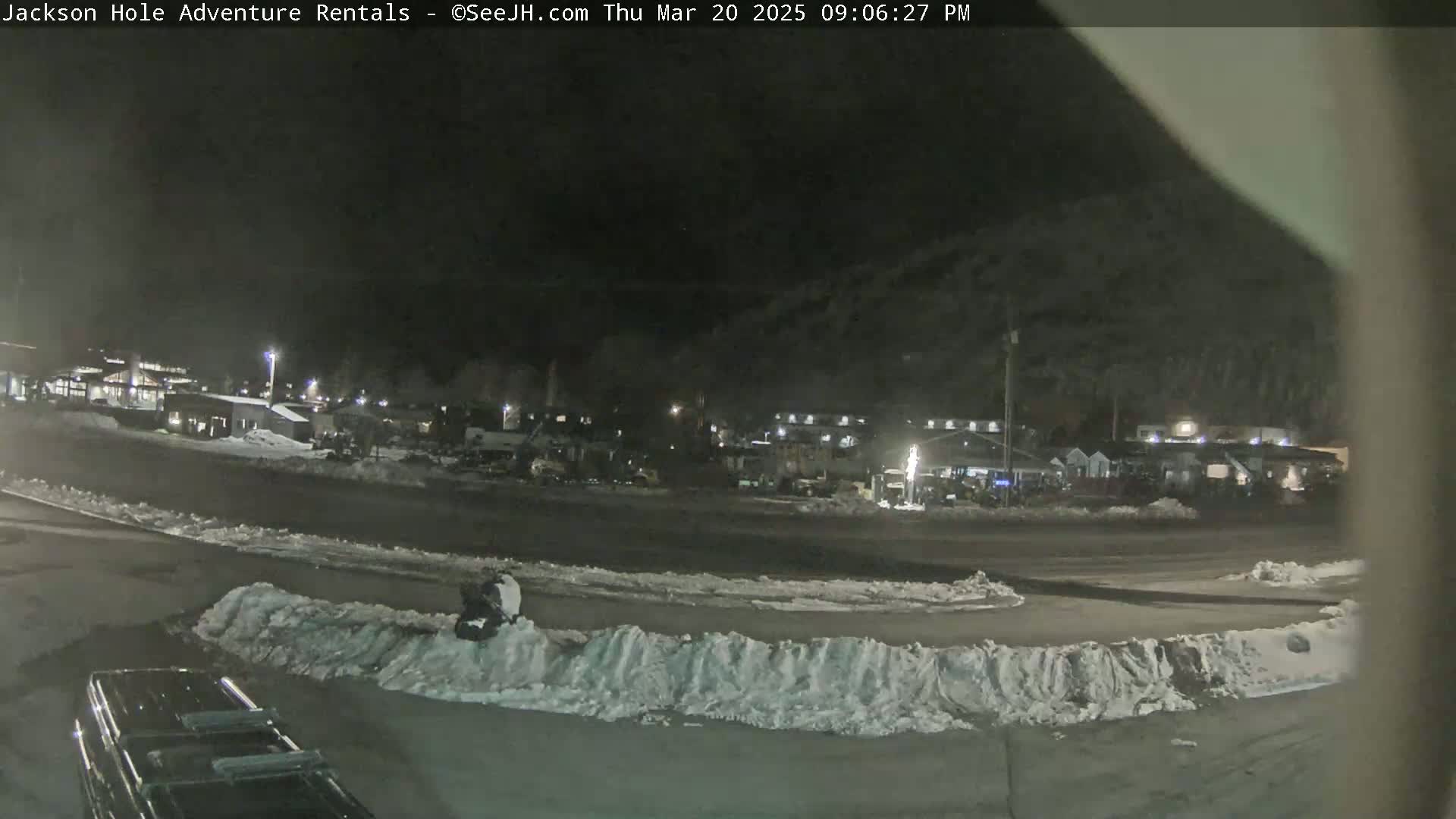 A nighttime view of a snow-covered parking lot and buildings in the distance under a dark sky.