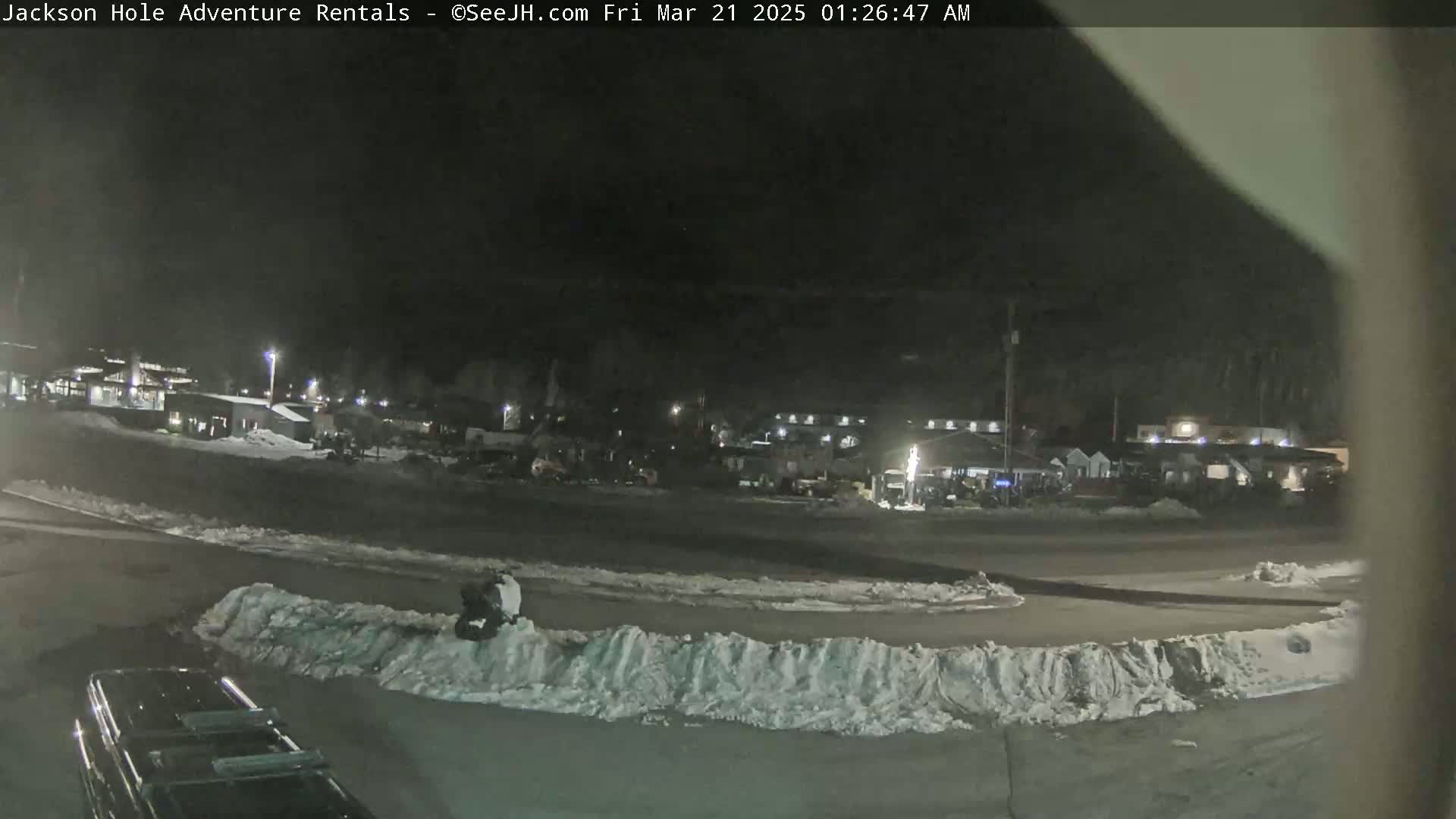 A nighttime scene shows a snow-covered parking lot with several buildings in the background under dark conditions.