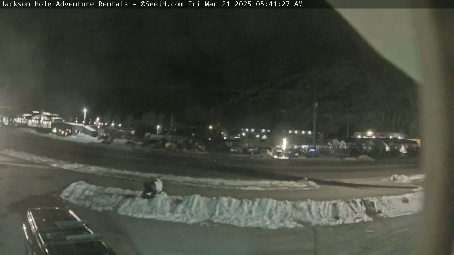 A nighttime, snow-dusted parking lot with several buildings and vehicles visible in the background.