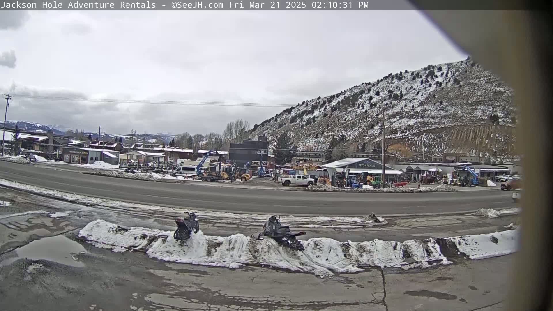 A snow-dusted hillside overlooks a street with parked vehicles, some snow on the ground, and two snowmobiles parked near a snowbank.