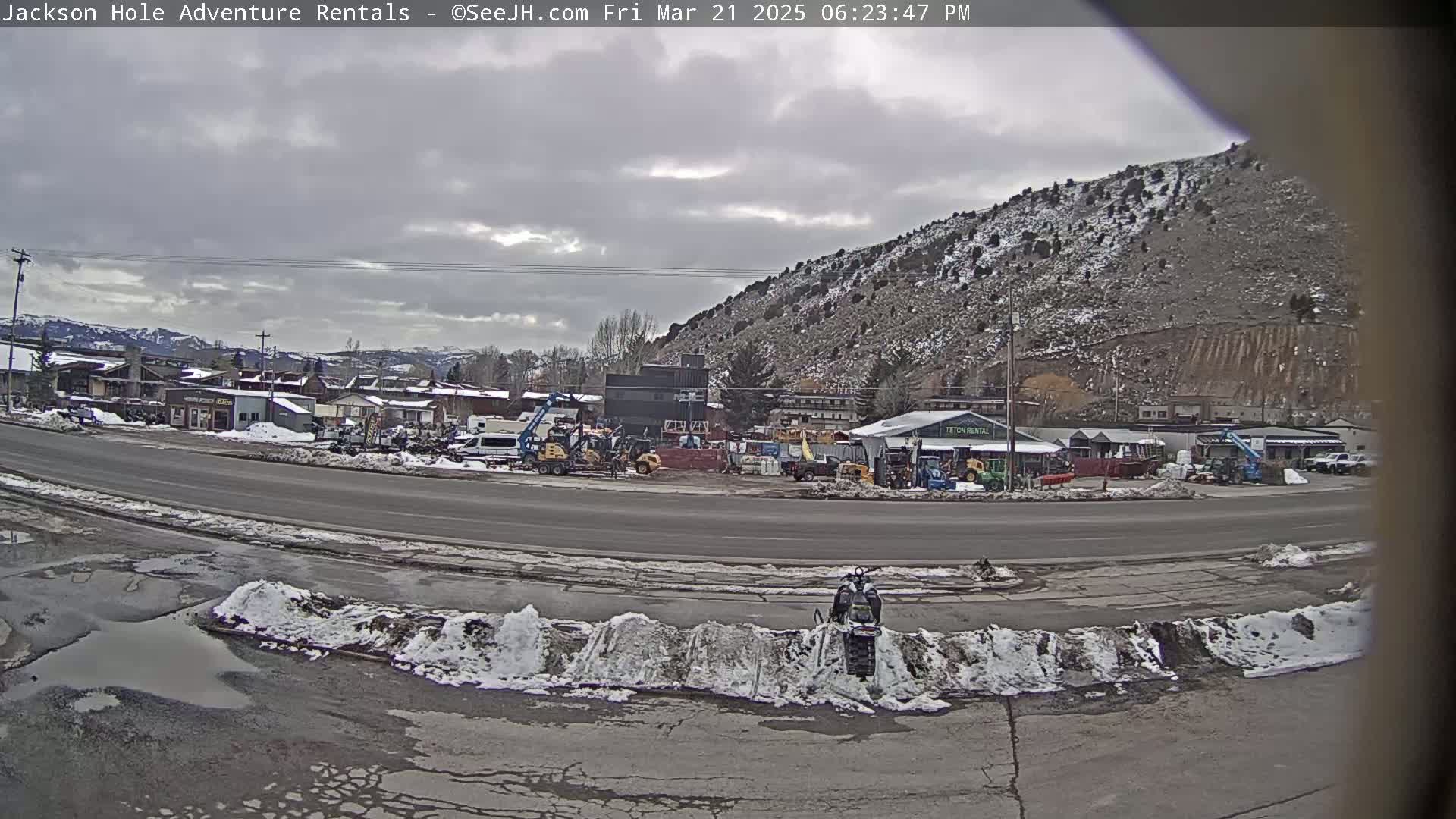 A snow-dusted mountainside overlooks a road with patchy snow and puddles, where various vehicles and equipment are parked near some buildings.