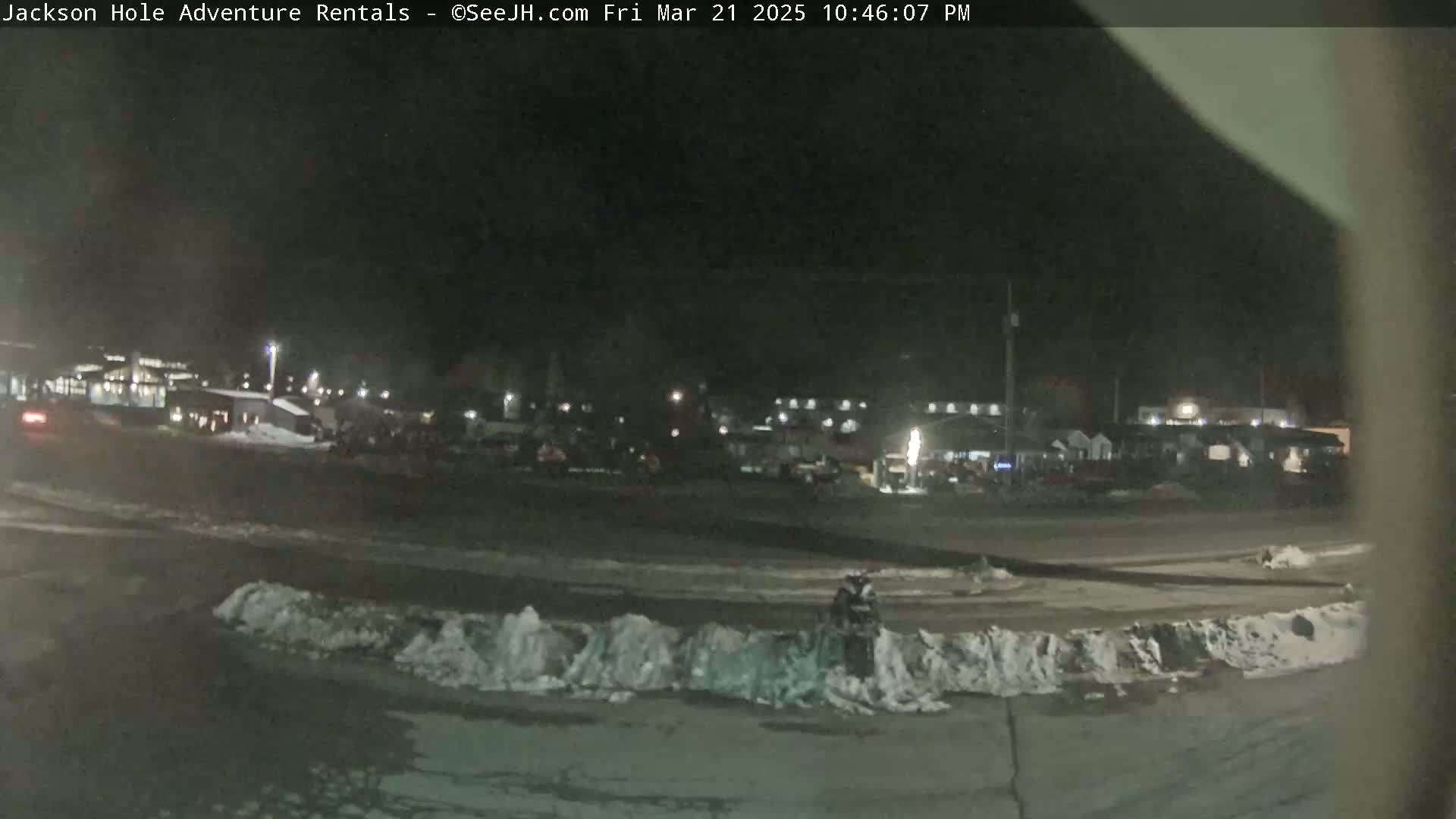 A nighttime, snow-covered parking lot with buildings and vehicles visible in the background under dark skies.
