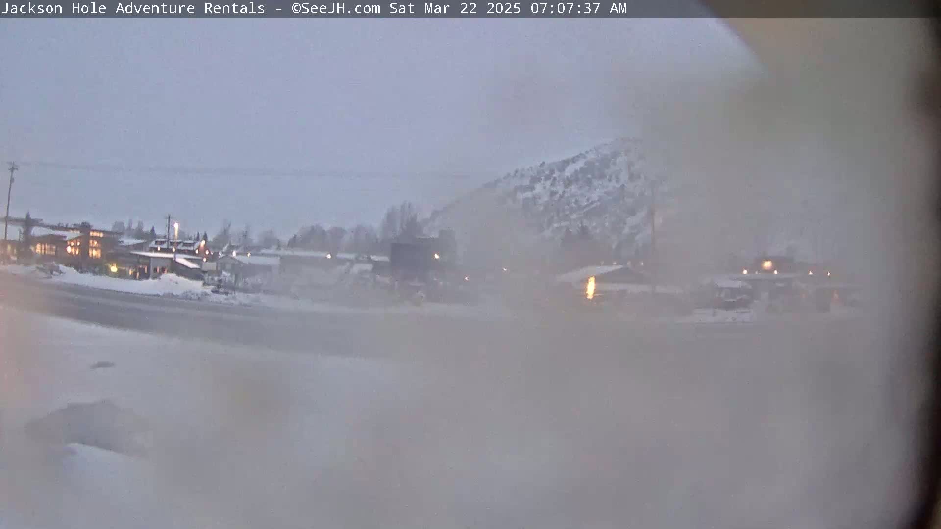 A hazy, snowy scene shows a cluster of buildings near a road and a snow-covered hillside under an overcast sky.