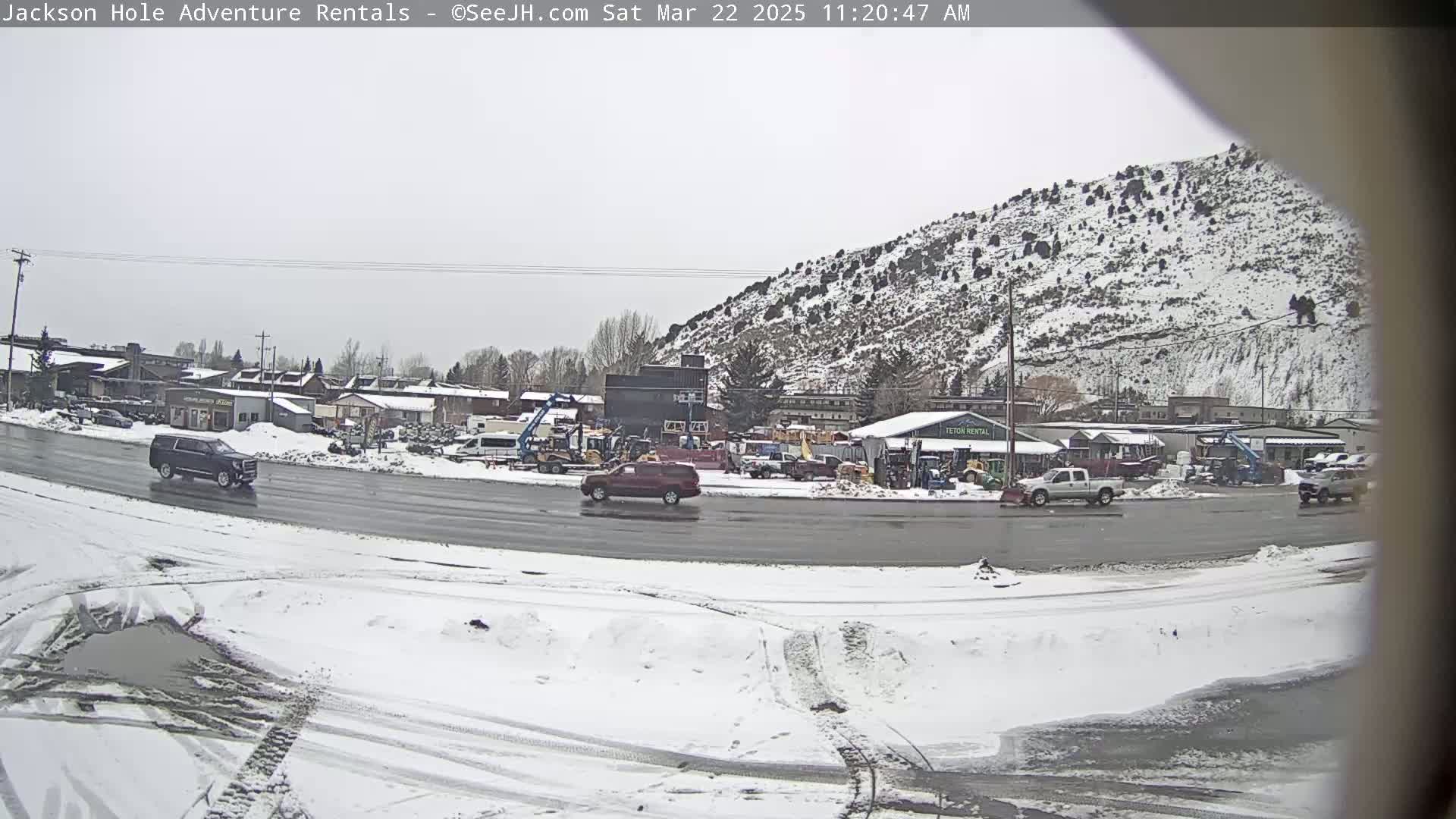 A snow-covered landscape shows a road with several vehicles and a background of buildings and a snow-dusted hill.