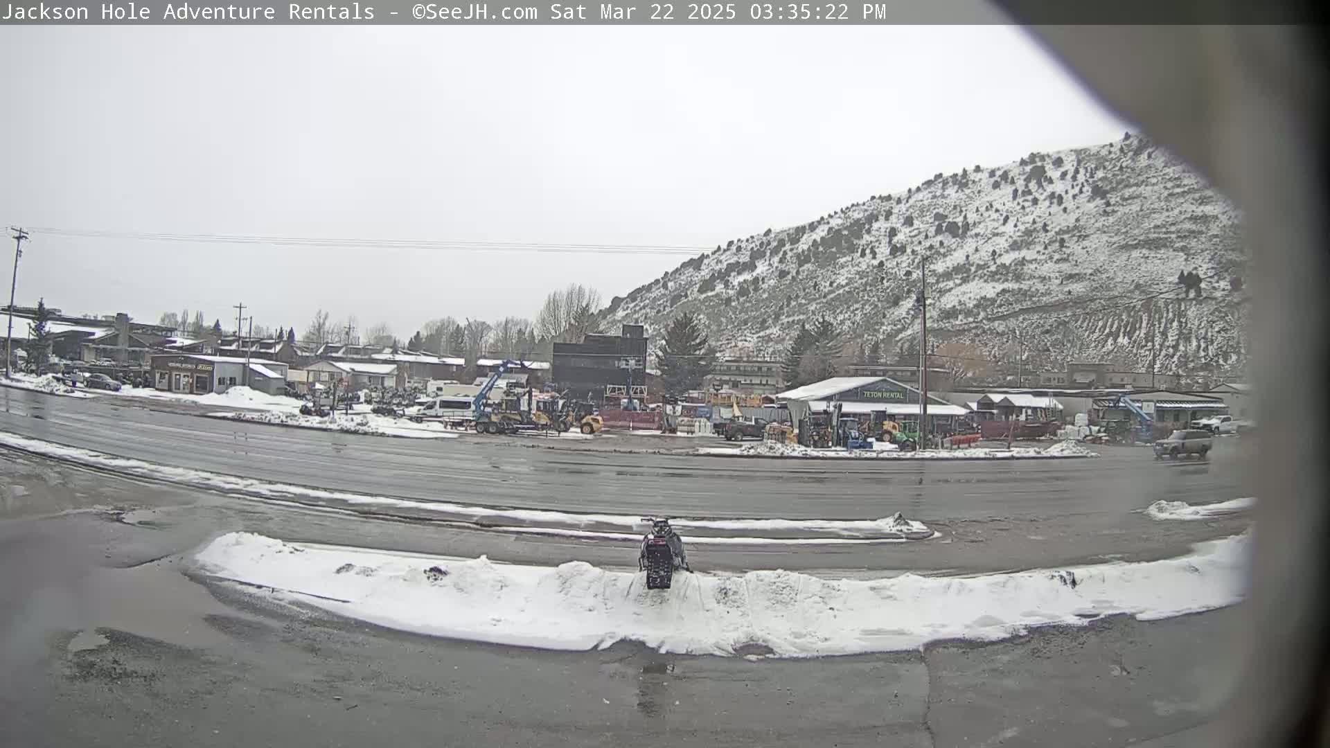 A snow-dusted mountainside overlooks a street scene with parked vehicles and construction equipment, under an overcast sky.