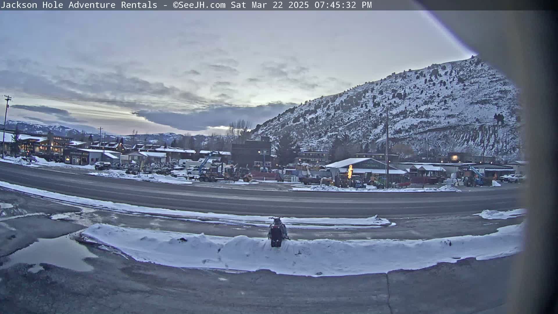 A snow-covered parking lot with a snowmobile, a road, and a town nestled beneath a snow-dusted mountain range under an overcast sky.