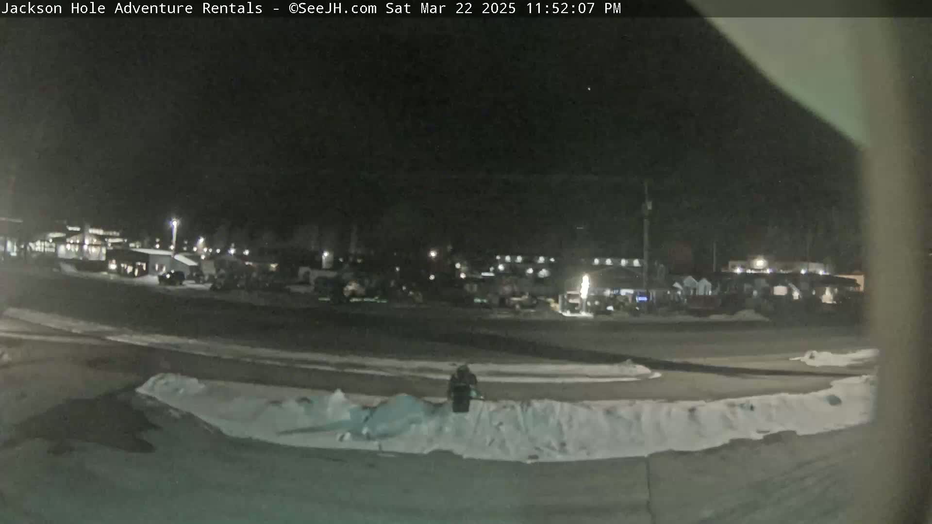 A nighttime, snow-covered parking lot with several buildings and vehicles visible in the background under a clear, dark sky.