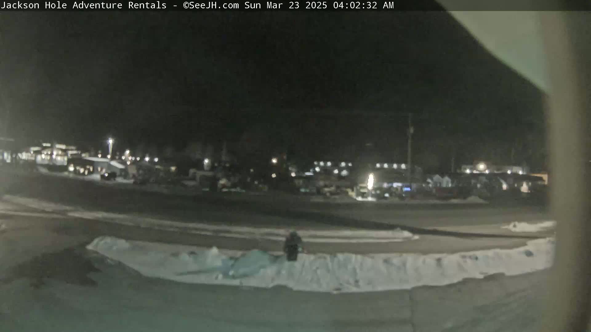 A nighttime, snow-covered parking lot with several vehicles and buildings in the background under dark conditions.
