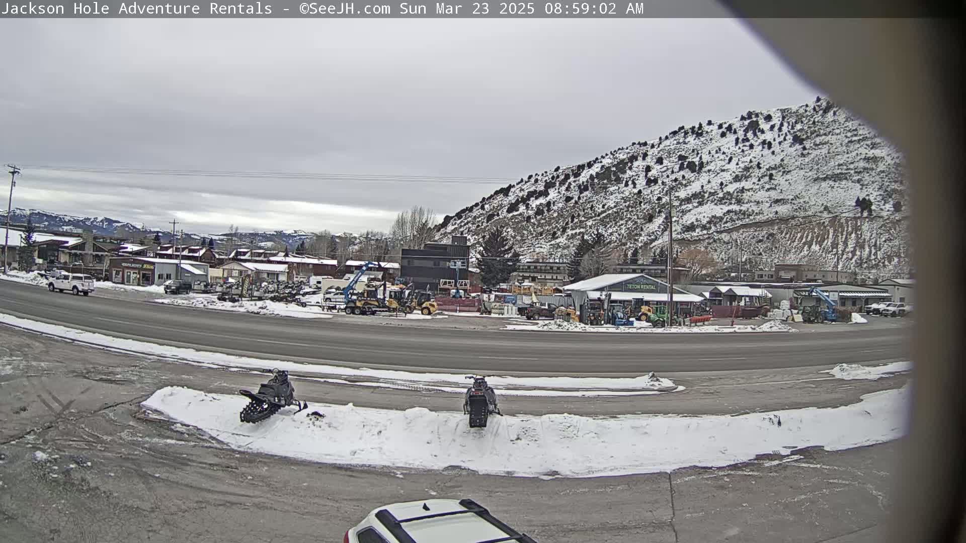 A snow-covered landscape shows a street with two snowmobiles parked and several buildings, including a rental business, against a backdrop of a snow-dusted mountain under an overcast sky.