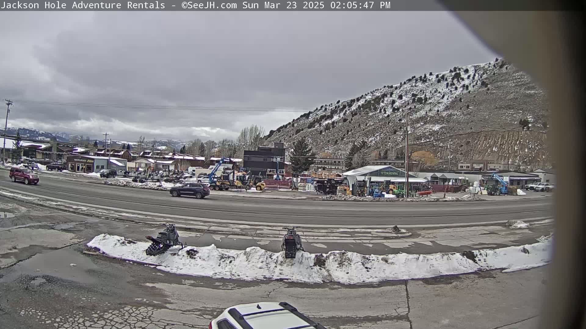 A cloudy, overcast day reveals a street scene with several vehicles, snow-covered ground, and a snow-dusted mountain in the background.