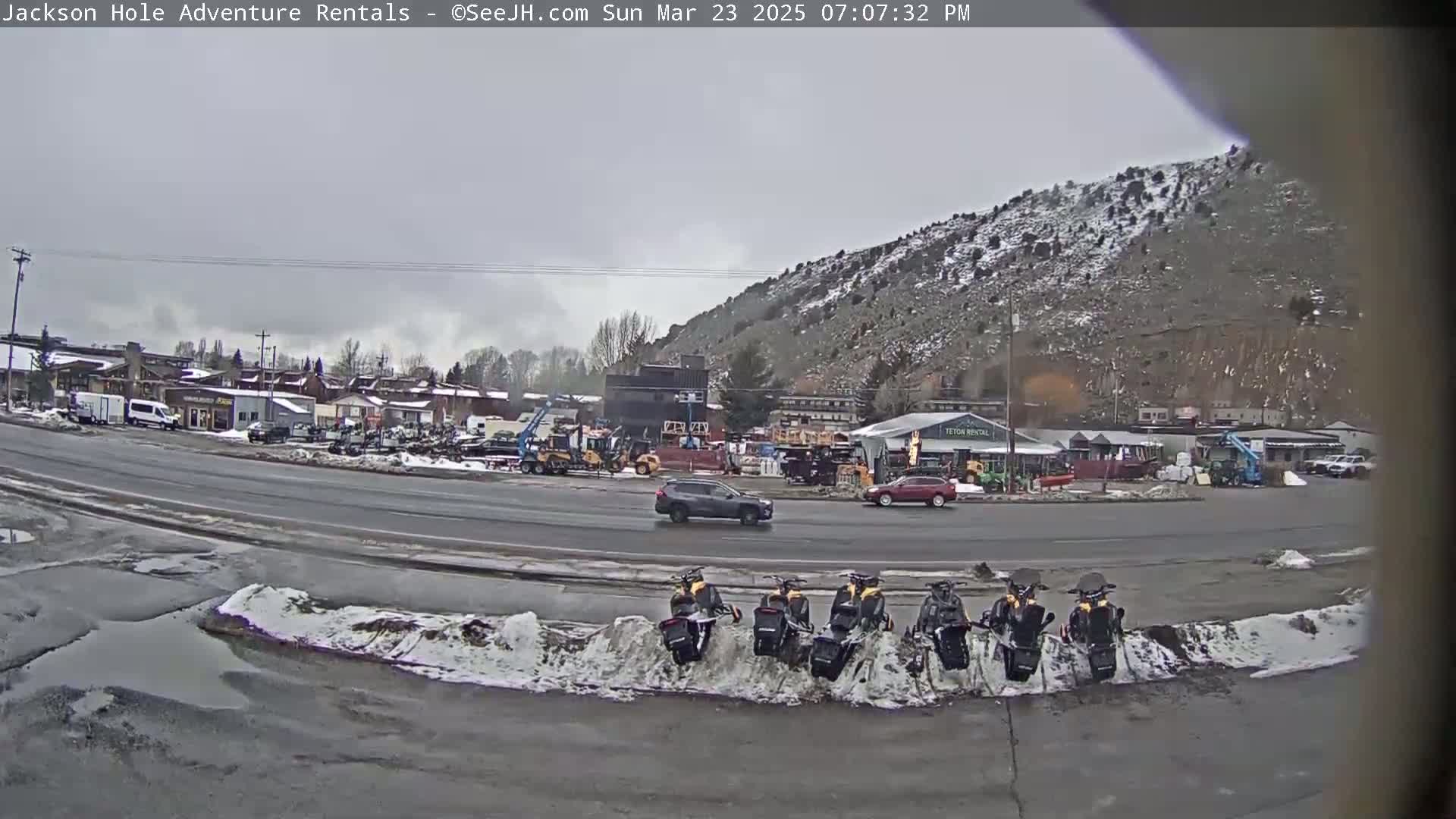 A gray overcast day shows a street scene with several vehicles parked and driving near a snow-dusted hill; several snowmobiles are parked along a snow bank beside the road.