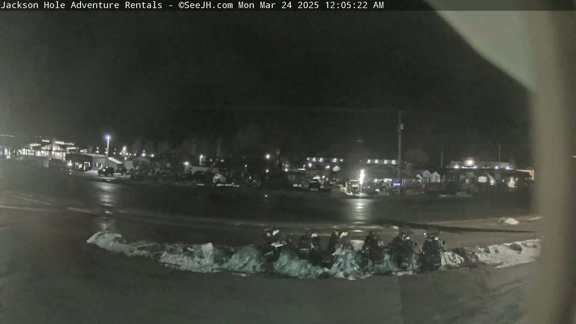A nighttime, snow-dusted parking lot with several snowmobiles parked near a building complex under a dark sky.